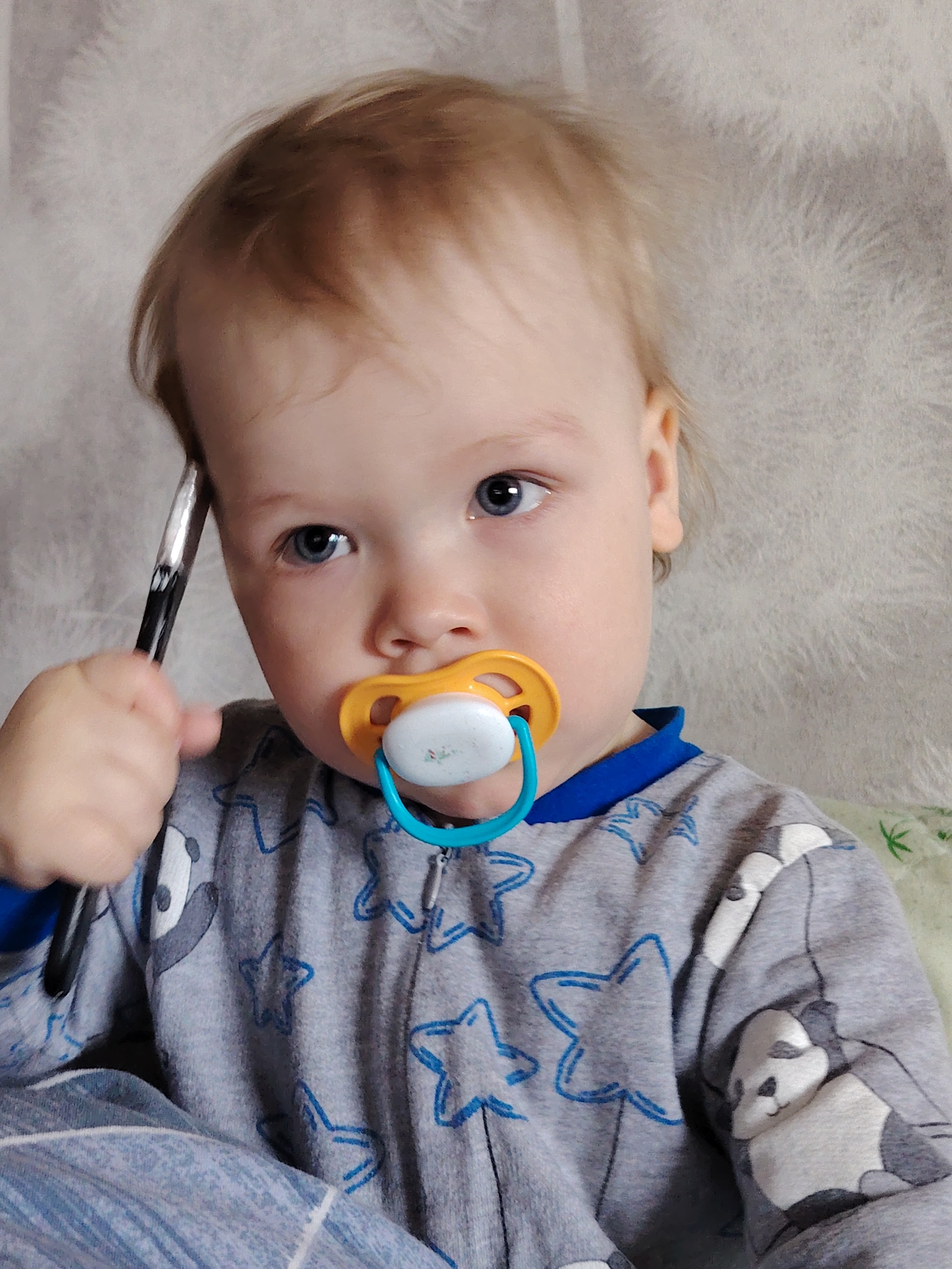 A young, light-skinned child with blond hair and blue eyes is shown from the chest up. The child is holding a black and silver pen against their head near the left temple. The child has a pacifier in their mouth with a yellow flange and a blue handle. They are wearing a gray pajama top with blue trim at the neck and blue star patterns. A panda bear design is on the lower right of the pajama top. The background is a light beige wall with a textured, possibly fur-like pattern. The image was taken in a building that is located in the city of Chaikovsky, Russia.