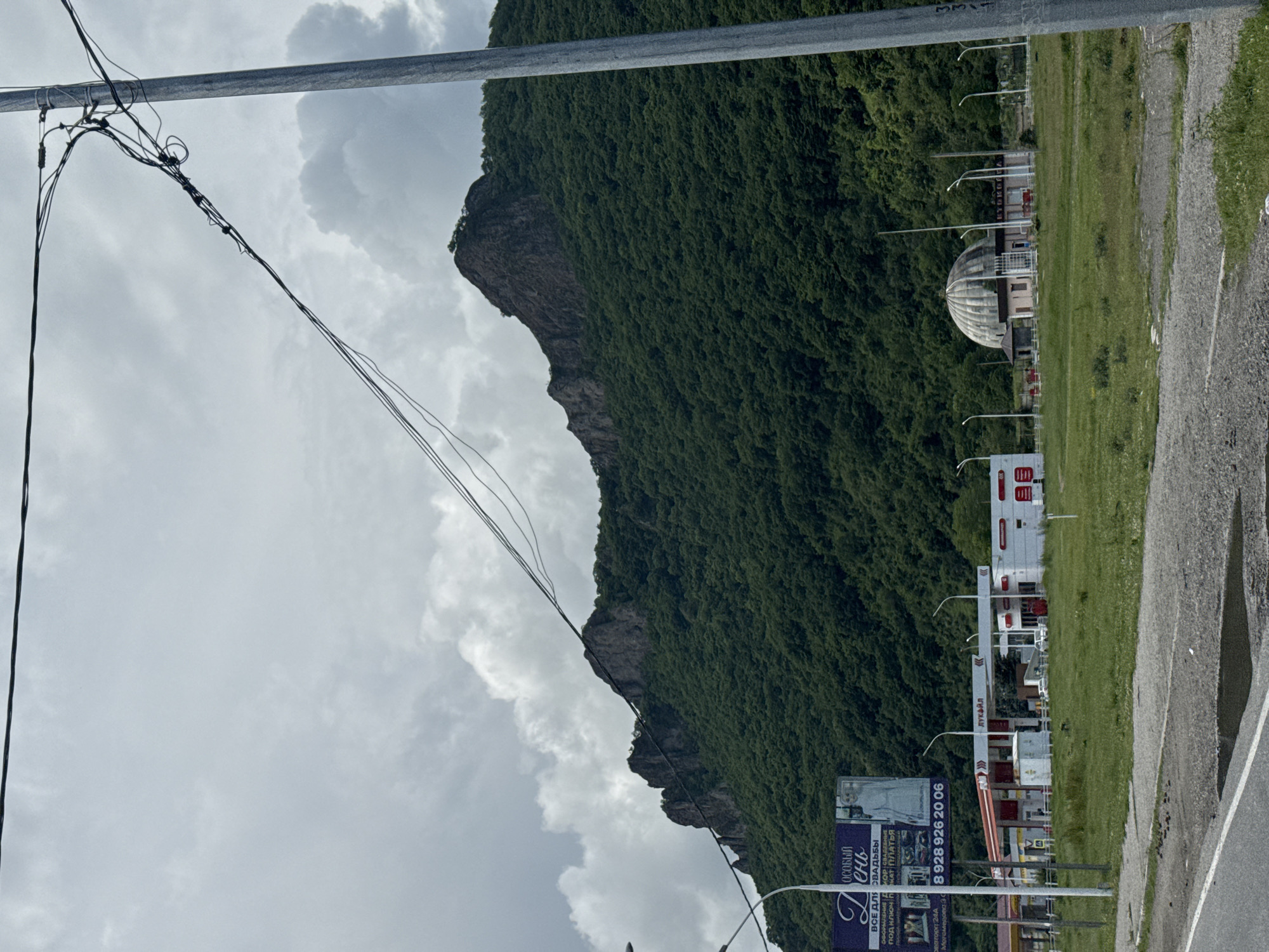 In Ordzhonikidzevsky, Russia, a desolate landscape unfolds under a heavily clouded sky. A rugged, tree-covered mountain dominates the background. In the foreground, a row of modern gas stations, including one with "LUKOIL" signage, stands alongside a distinctive, dome-shaped building. A large billboard, featuring an advertisement with text in Russian and an image of a woman, is also visible. Power lines crisscross the sky, and a tall utility pole stands on the right side of the frame. The scene is devoid of any visible people, and the overall atmosphere is one of quiet desolation.
