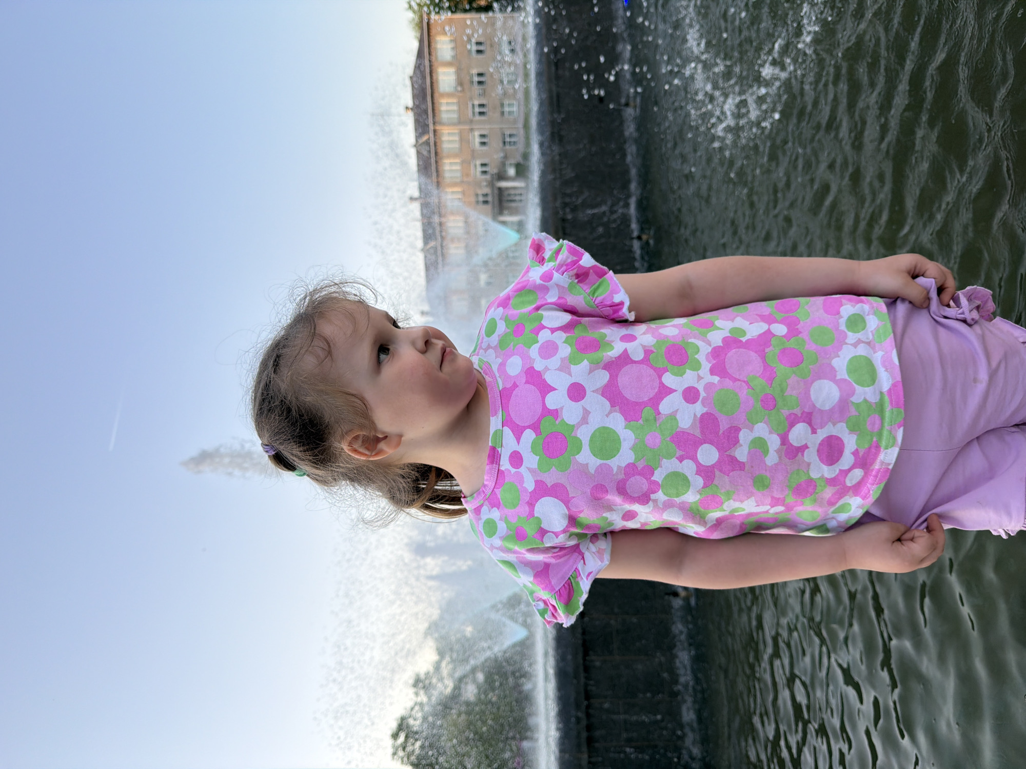 A young child with light brown hair, secured with small, colorful elastic bands, stands beside a fountain, gazing upward. She is dressed in a pink and white floral shirt and light purple pants. The fountain is actively spraying water upward, with a building visible in the background. The location is Chaikovsky, Russia. The child is at the edge of the fountain.