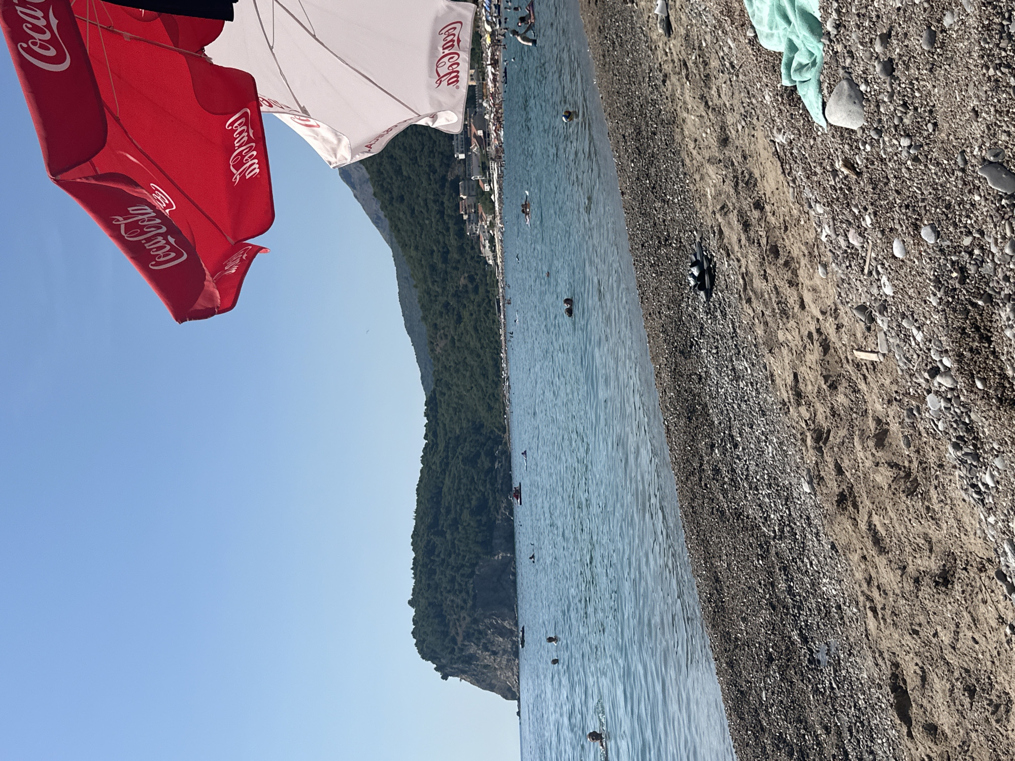 This is an eye-level, outdoor, medium shot of a pebbly beach leading to the Adriatic Sea. The water is a clear blue, with small waves and several people swimming and wading. A large, forested island with a rocky cliff face is visible in the distance, along with buildings and a densely packed beach with numerous sunbathers. Two large Coca-Cola branded beach umbrellas, one red and one white, are in the foreground, casting shadows on the sand. On the beach, a light blue towel and a pair of dark sandals are visible. The sky is clear and blue. This scene is located in the city of Đurovići, Montenegro.