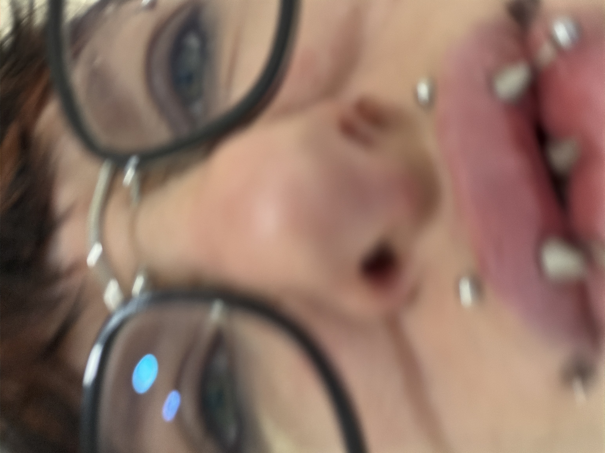 The image shows a close-up, distorted view of a person's face. The person is wearing glasses with black frames. The person's eyes are visible behind the lenses. The person has a nose piercing and multiple piercings on their tongue. The background is out of focus, making it impossible to determine the location in the city of Busmantsi, Bulgaria.
