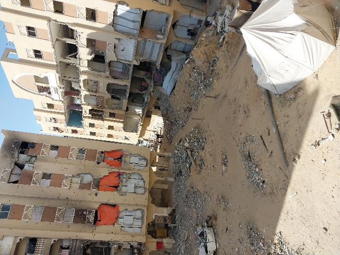 The image depicts a scene of extensive destruction in Khan Yunis, Palestinian Territory. Two severely damaged multi-story residential buildings dominate the background. The building on the left, taller and closer, exhibits numerous small impact holes across its light-colored facade, suggesting bullet or shrapnel damage. Multiple windows are shattered or missing, with some lower-level openings covered by bright orange tarpaulins. The upper section of this building shows charring and significant structural collapse. The building on the right also displays heavy damage, with large sections of its exterior walls missing, revealing exposed interiors, broken balconies, and debris hanging from openings.
The ground between and in front of the buildings is a sandy expanse heavily littered with rubble, concrete fragments, twisted metal, and other debris. In the midground, a white, burnt-out vehicle chassis is visible amidst the debris. In the lower right foreground, a white, hexagonal tent or temporary shelter is pitched on the sandy ground. A long, grey pipe or conduit lies horizontally across the lower midground. A few indistinct human figures are faintly discernible in the shadowy area at the base of the buildings, navigating the debris-filled environment. The sky above is clear and blue.