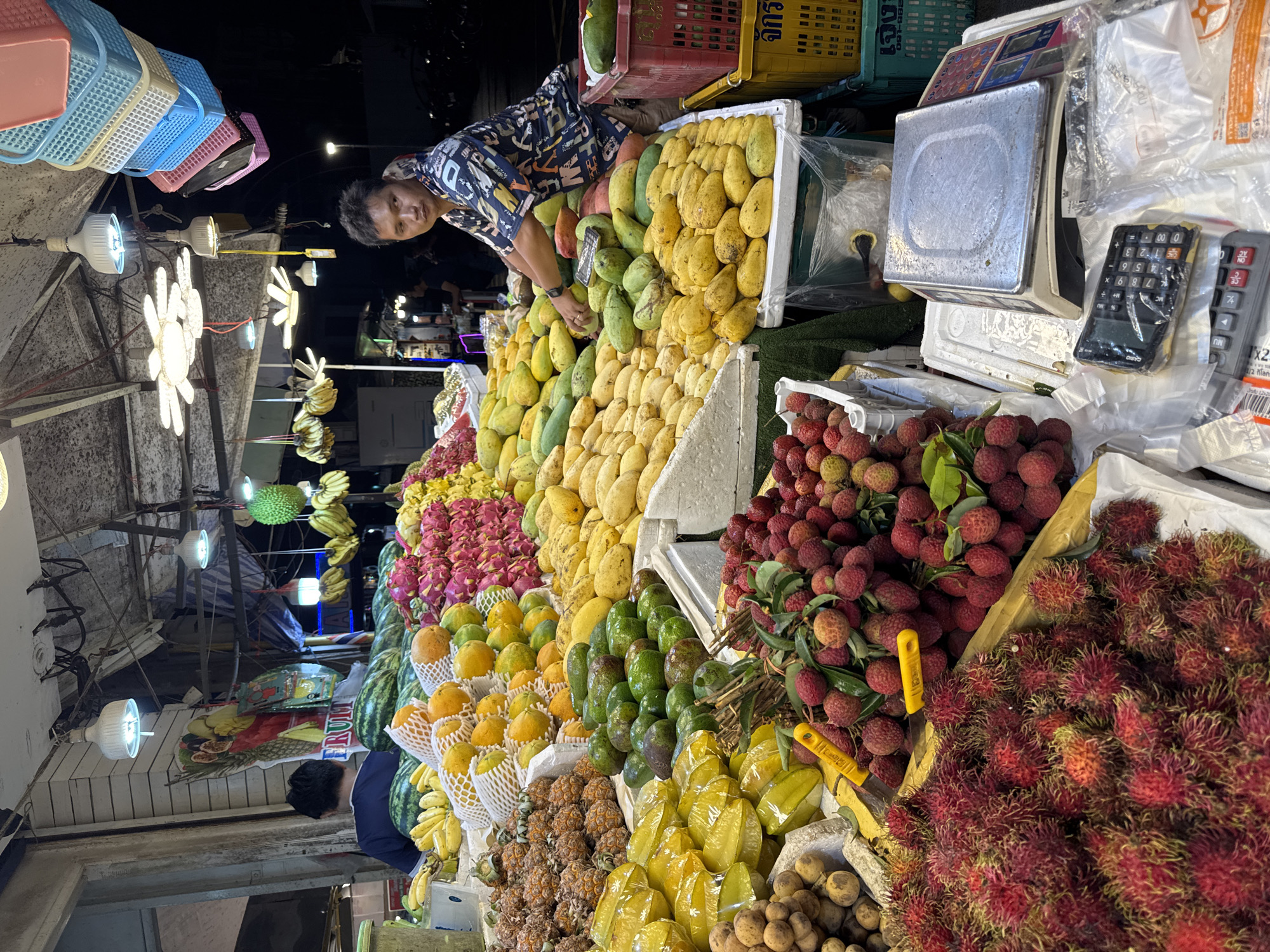 A fruit stand in Karon, Thailand is depicted at night. The stand is filled with an abundance of fresh fruit, including rambutan, lychee, starfruit, mangoes, avocados, papayas, dragon fruit, pineapples, bananas, and watermelons. A vendor, a man with short dark hair, stands behind the fruit, wearing a patterned shirt. He is looking towards the viewer and appears to be interacting with the fruit. Various objects are present at the stand, such as a scale, calculators, and plastic baskets. The scene is lit by overhead lights, casting shadows and illuminating the colorful display of produce. The background suggests an outdoor market setting with other stalls and possibly a street visible.