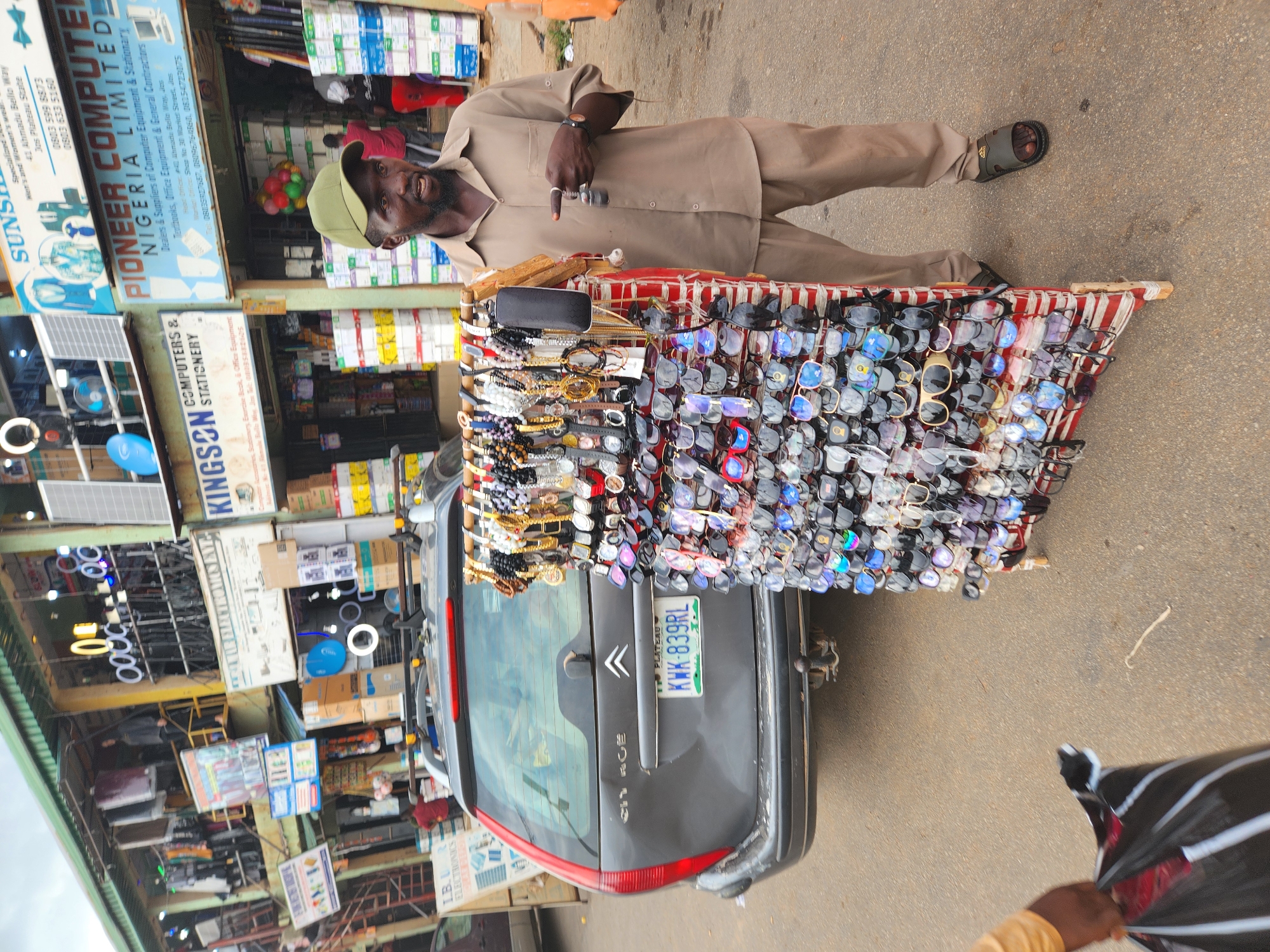 The image shows a street vendor in Jos, Plateau State, Nigeria, standing beside a car laden with sunglasses and watches. The man, wearing a green cap and khaki clothing, stands next to a grey Citroen C4 with the license plate KWK-839RL. The vehicle is covered with a red and white striped stand displaying numerous pairs of sunglasses and wristwatches. In the background, storefronts are visible, including "KINGSON COMPUTERS & STATIONERY" and "PIONEER COMPUTER NIGERIA LIMITED," indicating the location is likely a commercial area. A sign for "SUNS" is also visible. The street surface appears to be paved. A partially visible person is holding a black bag in the foreground.