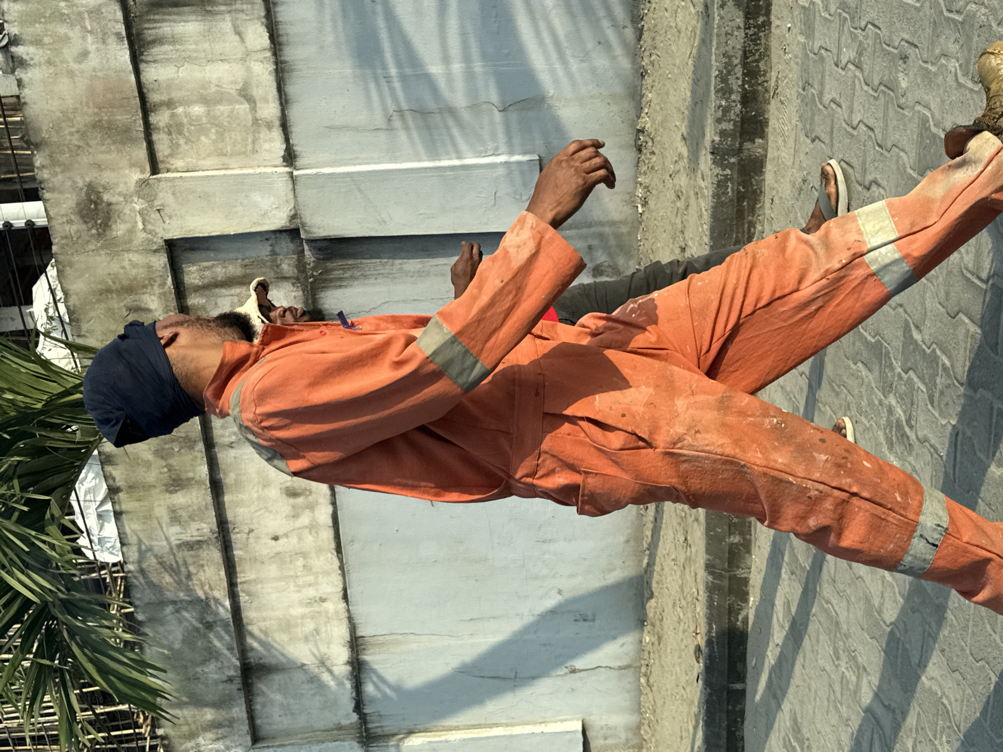 An eye-level, medium shot captures two individuals walking outdoors in Igboefon, Nigeria. The primary subject, a person wearing an orange jumpsuit and a dark blue head covering, is positioned in the foreground, walking from left to right across the frame. Their jumpsuit is stained and appears worn, with reflective grey bands around the lower legs and upper arms. To the right of the main subject, a second individual is partially visible, also walking away from the camera, wearing a light-colored hat and a dark shirt. They are walking on a paved surface composed of interlocking grey stones. Behind the individuals is a textured, grey concrete wall with visible staining and grime. Above the wall, lush green palm fronds extend into the upper left corner of the image. The lighting suggests it is daytime, with shadows cast on the ground.
