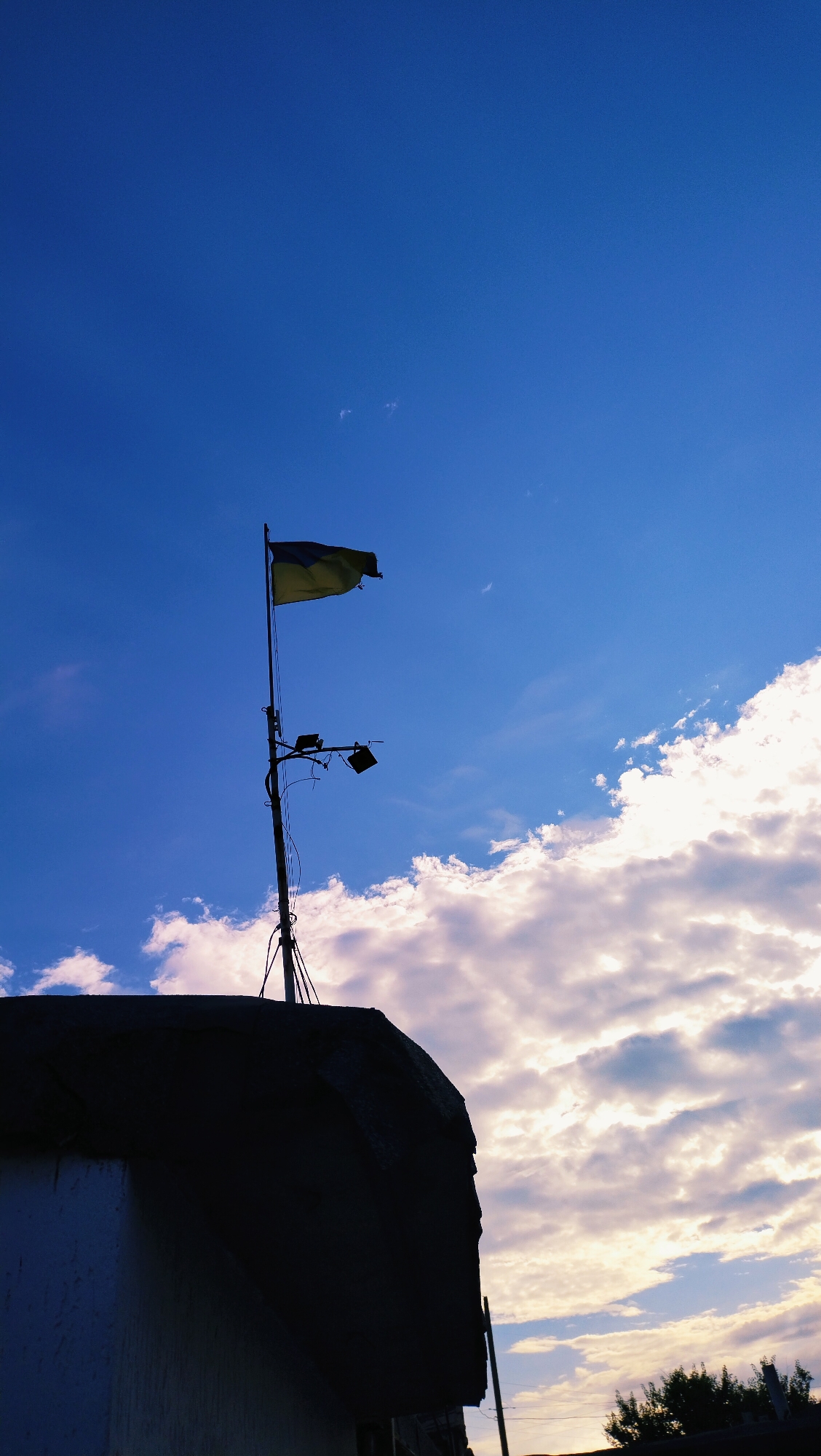 In Horishni Plavni, Ukraine, a Ukrainian flag, yellow and blue, flutters atop a metal pole. The pole is adorned with two black floodlights. The structure supporting the pole is silhouetted against a vast expanse of blue sky, punctuated by scattered white clouds. The silhouette also reveals the edge of a building with a dark, textured roof.