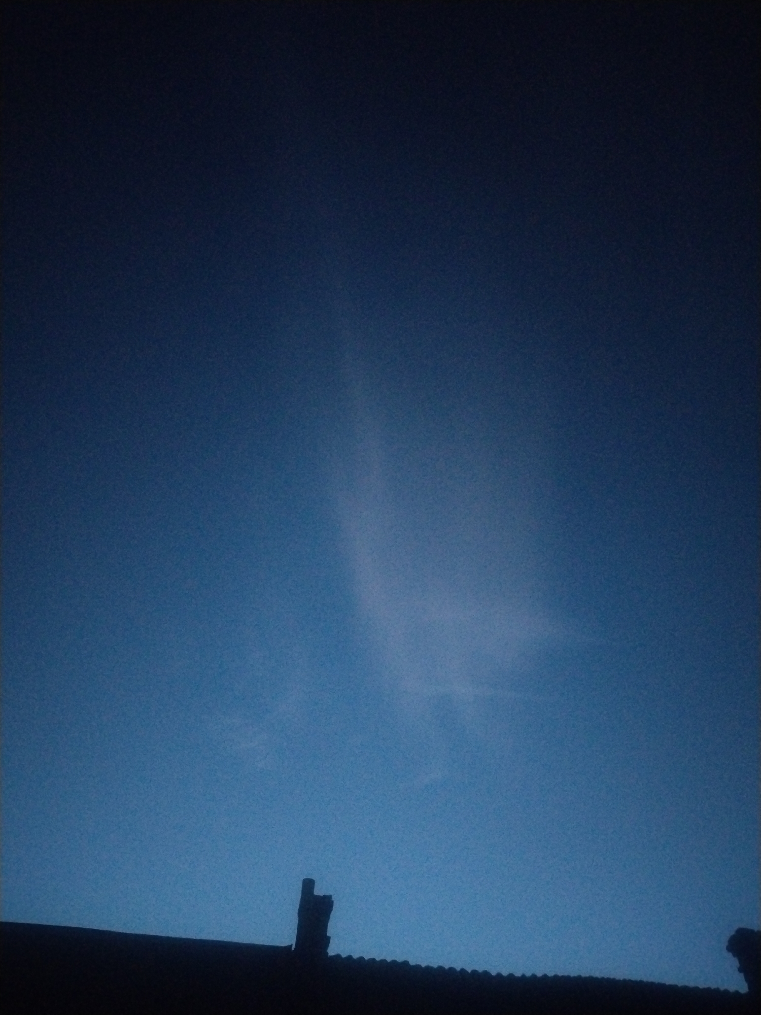 The image shows a dark blue sky with a faint cloud formation. In the foreground, the silhouette of a rooftop with a chimney is visible. The setting appears to be at dusk or dawn, given the low light conditions. The scene is located in Unknown, Unknown. There are no people or discernible actions taking place.