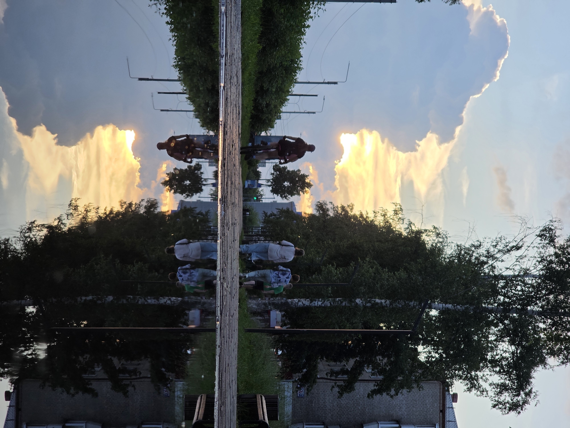 In Chaikovsky, Russia, at dusk, a cyclist rides away from the viewer on a path, their silhouette framed by a dramatic sky filled with illuminated clouds and the setting sun. The path, paved with cobblestones, is bordered by a grassy area and trees on the right, and lush green bushes and streetlights on the left. In the foreground, a large puddle reflects the scene, including the cyclist, the sky, trees, and a multi-story brick building visible in the background. Three figures walk along the path in the middle ground, also reflected in the puddle. The overall atmosphere is serene, with the warm light of the setting sun casting long shadows.