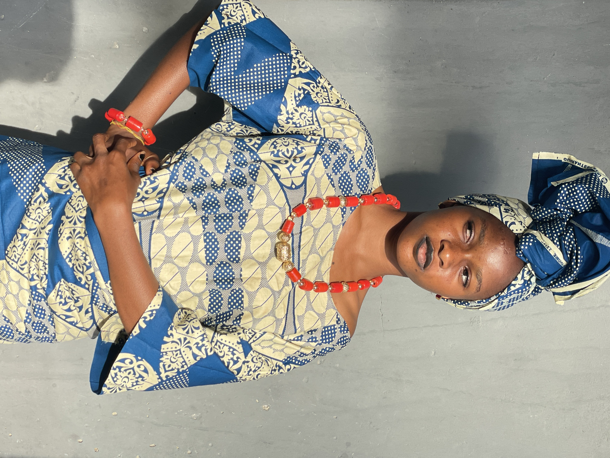 The image features a woman in traditional Nigerian attire, possibly in Badagry, Nigeria. She is wearing a blue and beige patterned top, a matching head wrap, and a red beaded necklace and bracelet. She is posing on a gray surface that appears to be concrete. The lighting indicates it might be outdoors. Her expression is neutral, and she is looking slightly upwards. Her hands are clasped in front of her.