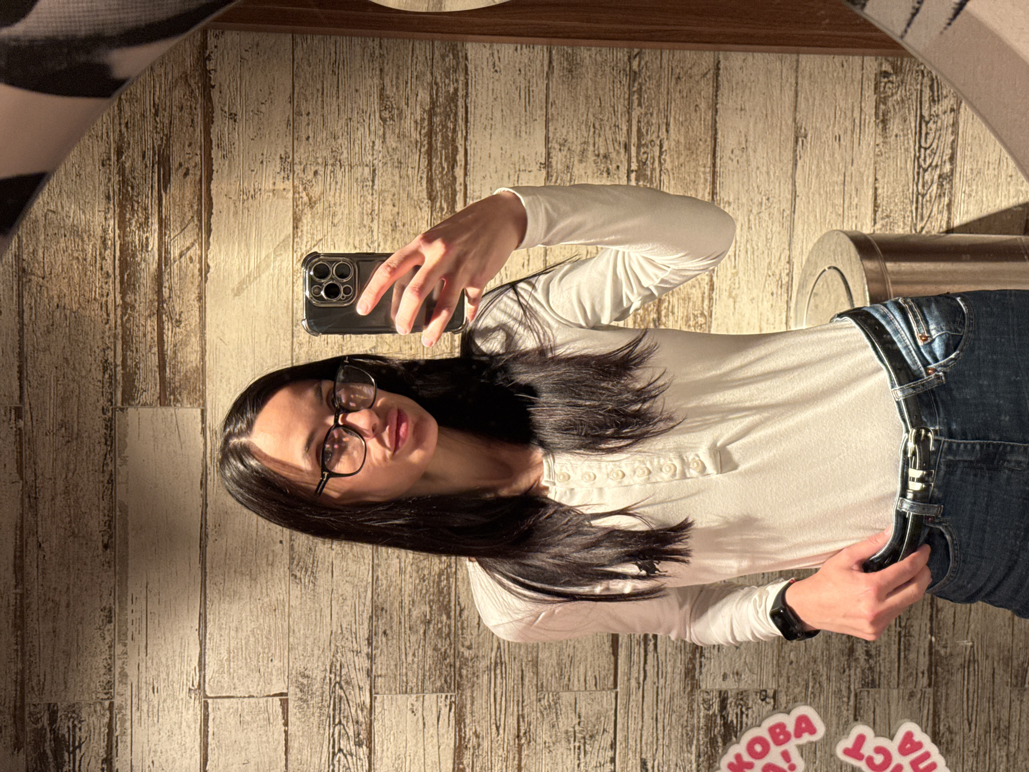 A woman is taking a selfie in a mirror. She is wearing glasses, a long-sleeved white shirt, and jeans with a black belt. Her dark hair is long and loose. She is holding her phone up with her right hand, and her left hand is near her waist. A stainless steel trash can is visible in the background. The background also features what appears to be a wooden-textured wall. Pink stickers with letters on them are on the floor near the bottom of the image. The location is likely inside a room, but the specific location in Sofia, Bulgaria, is not evident from the image alone.