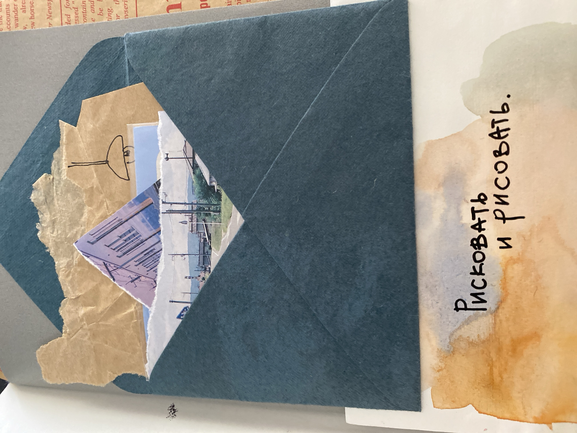 The image displays a collage within an open blue envelope. Inside the envelope are several cut-out pieces of paper including an old newspaper clipping, a torn piece of brown paper with a simple line drawing of what appears to be a street light, and a partially visible photograph showing buildings and power lines. To the right of the envelope, on a white background, is a watercolor stain in shades of brown and blue. Written vertically in black ink in Russian, the phrase "Рисковать и рисовать" (Riskovat' i risovat') appears, which translates to "To risk and to draw" or "To venture and to paint."
The location in the city of Lâm Viên - Đà Lạt, Vietnam, is not discernible from the visual information provided.