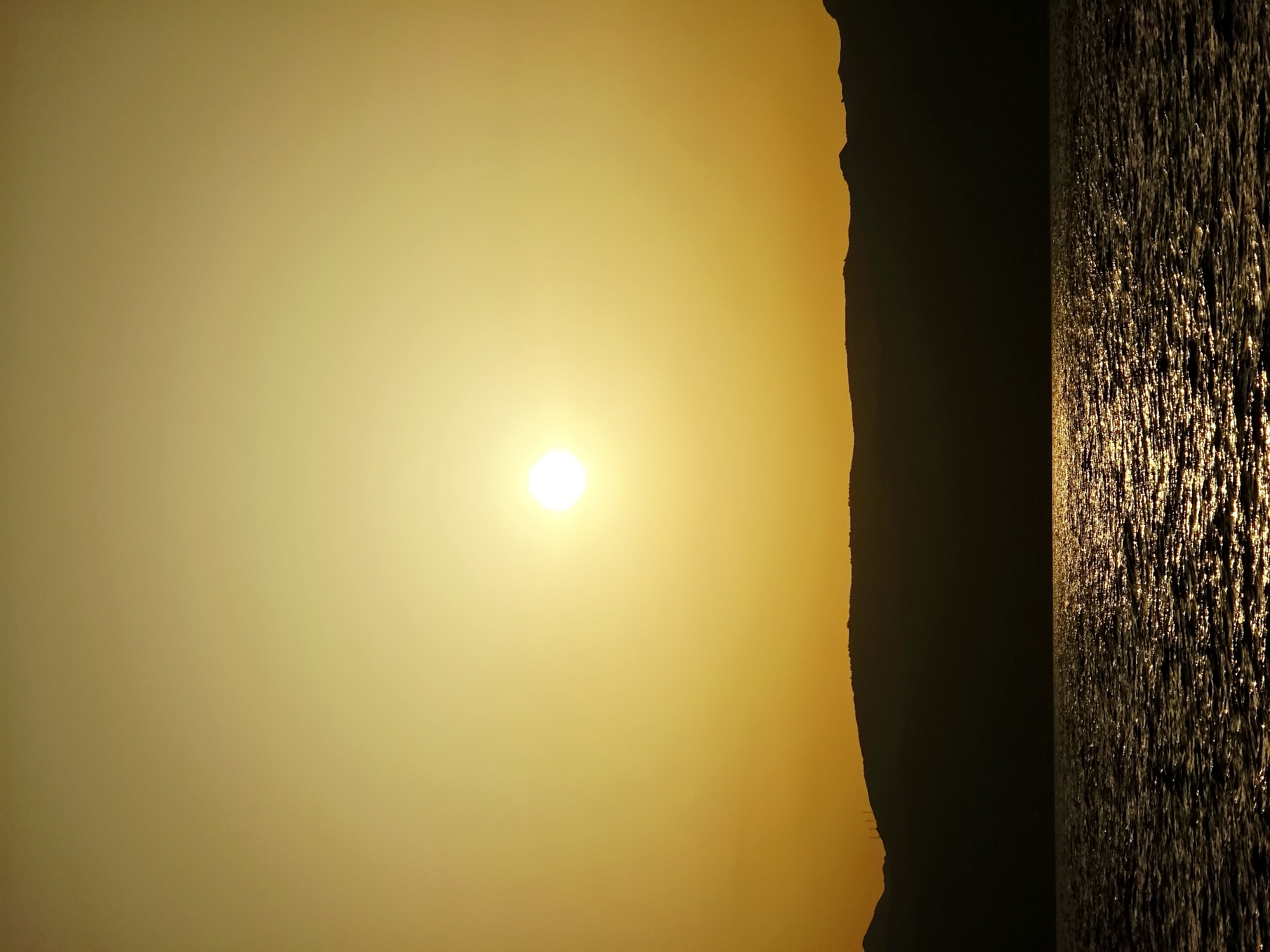 A vertical, silhouetted landscape photograph captures a sunset over the Aegean Sea, likely near the Heraklion Municipal Unit of Crete, Greece, given the characteristic coastal mountain range and clear, vast sky. The sun, a bright white disc, hangs low in the sky, radiating a warm, golden-yellow light that saturates the upper portion of the image. Below the sun, a dark, undulating silhouette of a mountain range stretches across the frame, its peaks and ridges sharply defined against the luminous sky. The lower half of the image is dominated by the sea, its surface rippling with dark, choppy waves. The setting sun casts a shimmering, golden reflection onto the water, creating a path of light that leads towards the viewer. No people or specific structures are discernible in the silhouette, suggesting a natural, unpopulated coastal scene. The overall impression is one of natural beauty and tranquility at the close of day.
