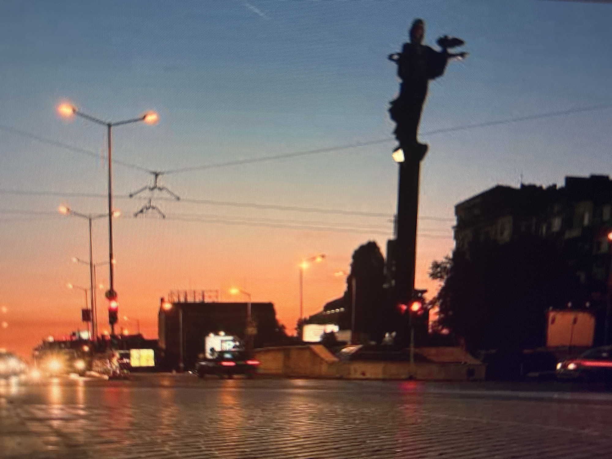 The image captures a street scene at sunset in Sofia, Bulgaria, dominated by the Statue of Sveta Sofia. The statue, silhouetted against the orange and blue sky, stands prominently on a tall pillar. Streetlights are visible along the road, as are traffic lights, which are red. Cars with headlights and taillights are moving on the wet cobblestone road, reflecting the ambient light. Buildings form a backdrop to the statue and street activity. Overhead power lines cross the sky.