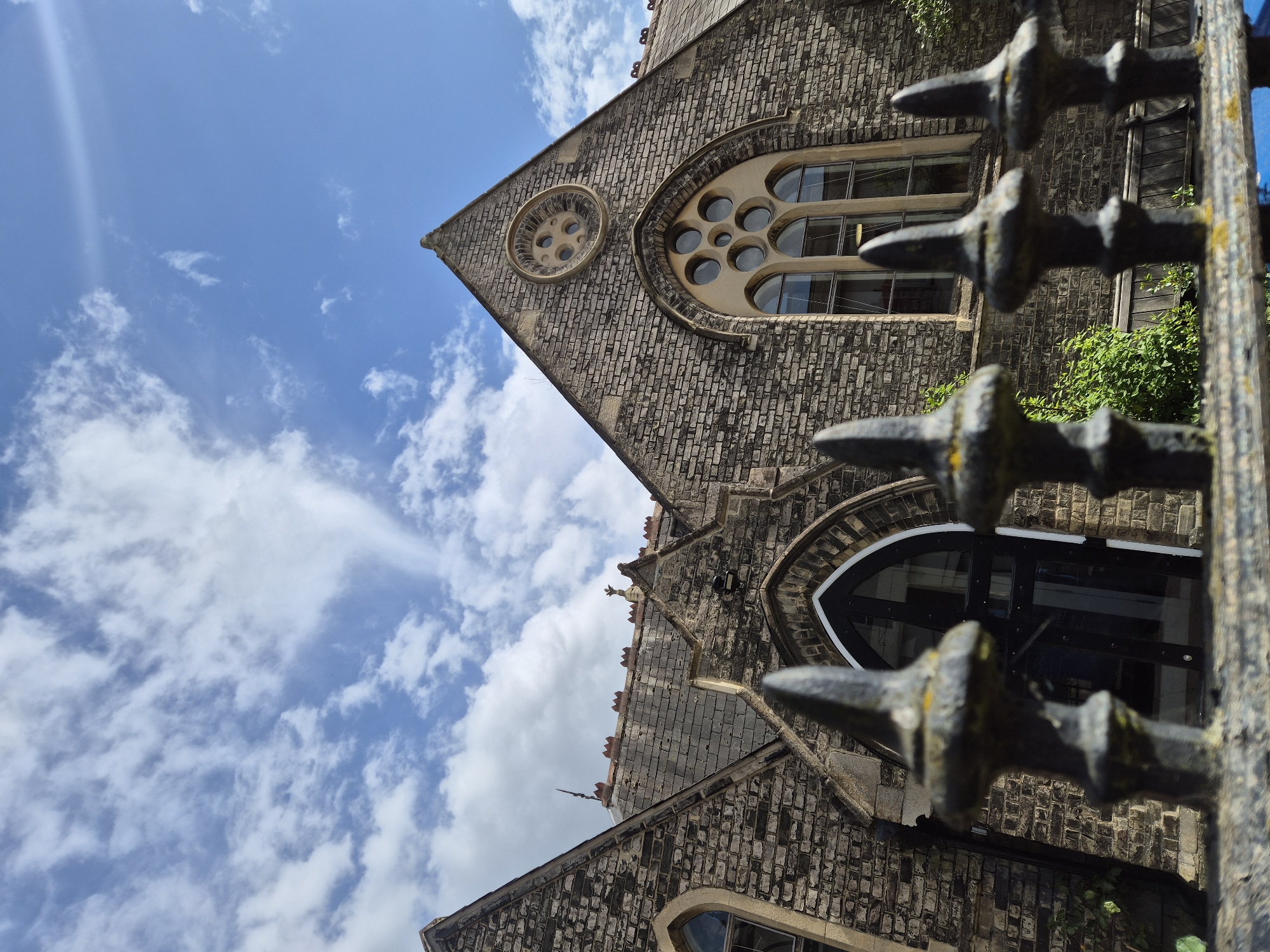 The image shows a building in West Suffolk, United Kingdom, viewed from a low angle through a metal fence. The building appears to be a stone structure, possibly a church or similar historical building, featuring arched windows, circular decorative elements, and a steeply pitched roof. The sky is visible in the background, with scattered clouds and a patch of blue. The metal fence, which is partially in focus, has decorative spikes on the top.