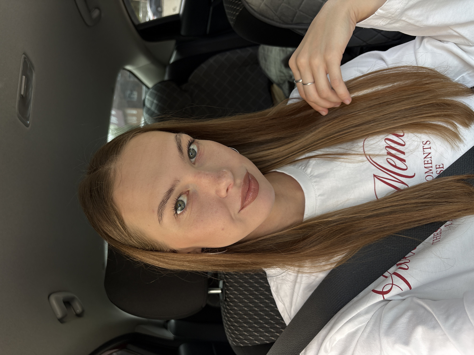 A young woman with long, straight, light brown hair is sitting in the passenger seat of a car. She is wearing a white long-sleeved shirt with red lettering. The car appears to be parked, as indicated by the safety belt being worn and the indoor lighting. The background shows the interior of the car, with dark seats and headrests. The woman is looking at the camera and smiling slightly. She has light green eyes and is wearing subtle makeup, including lipstick. A silver ring is visible on her right hand. The overall lighting suggests it is daytime. The location is not specified in the image.
