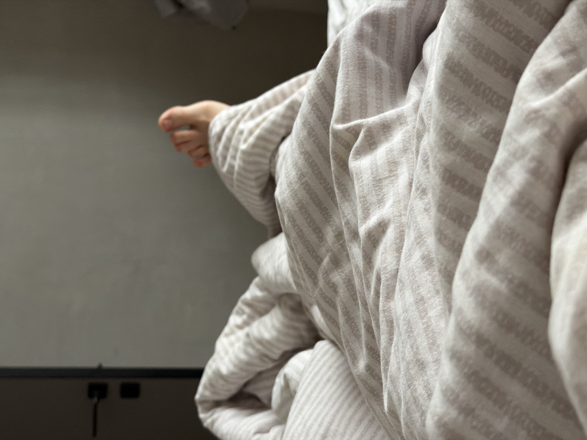 This is a close-up, low-angle shot of a person's legs under a striped duvet in a bedroom. The left side of the image shows a portion of a door with a dark handle and a black square detail, possibly a lock or a light switch. The background is a plain, neutral-colored wall. The duvet is light-colored with thin, darker striped patterns. A single foot, with visible toes, protrudes from under the duvet on the right side of the frame. The lighting appears natural, suggesting it may be daytime. The context of Chaikovsky, Russia, is not evident from the visual information in the image.