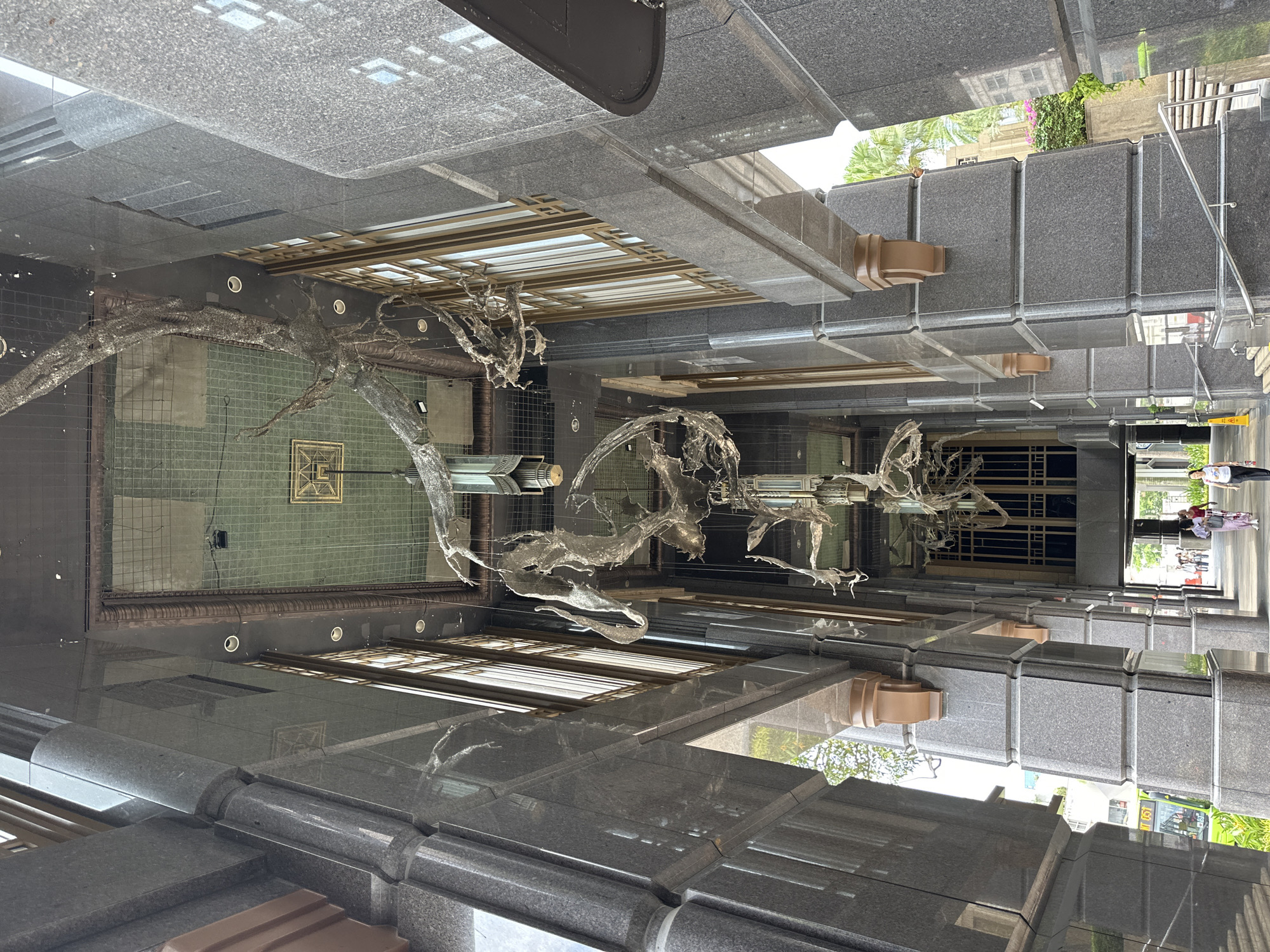 The image shows an interior view looking upwards of a covered walkway in Singapore, likely part of a commercial building. Large, dark granite pillars flank the walkway, with ornate brown brackets supporting the upper structure. Hanging from the ceiling are several large, abstract sculptures made of a metallic, mesh-like material that resembles draped fabric or organic forms. The ceiling itself is a grid of green tiles with some lighting fixtures visible. Tall windows with wooden frames line the upper portion of the walkway. Sunlight filters in from the open ends of the walkway, illuminating a path where several people are walking. In the distance, a green bus and tropical foliage are visible, suggesting an outdoor environment beyond the walkway. The perspective is from ground level, looking up and into the distance.