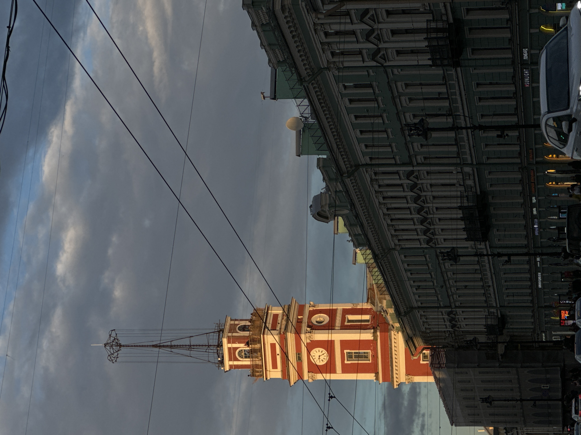 The image captures a street view in Saint Petersburg, Russia, featuring a prominent red and white clock tower. The tower, adorned with a clock face and architectural details, is a central element of the composition. Adjacent to the tower is a long, multi-story building with a pale green facade, characterized by numerous windows and classical architectural elements. The sky above is partly cloudy, with dramatic lighting suggesting either sunrise or sunset. Overhead, a network of power lines traverses the frame. In the foreground, a street is visible with several cars and pedestrians, indicating an urban setting. The scene is illuminated by the warm glow of the setting or rising sun, casting shadows and highlighting the textures of the buildings.