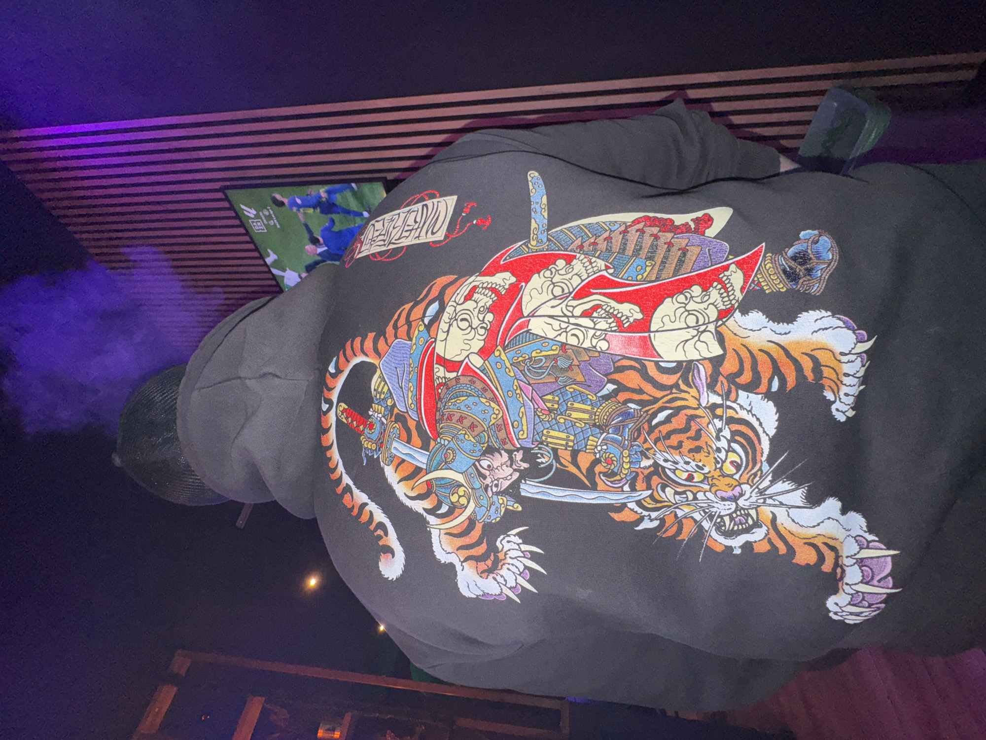 A person wearing a black hoodie with a colorful design of a samurai riding a tiger is seen indoors. The hoodie's design features intricate details and vibrant colors. The person is wearing a black cap, and smoke is faintly visible near their head. In the background, there's a TV screen displaying what appears to be a soccer match. The scene is lit with purple light, and a wooden structure is visible at the bottom of the frame. I cannot confirm if the location is Barcelona, Spain.
