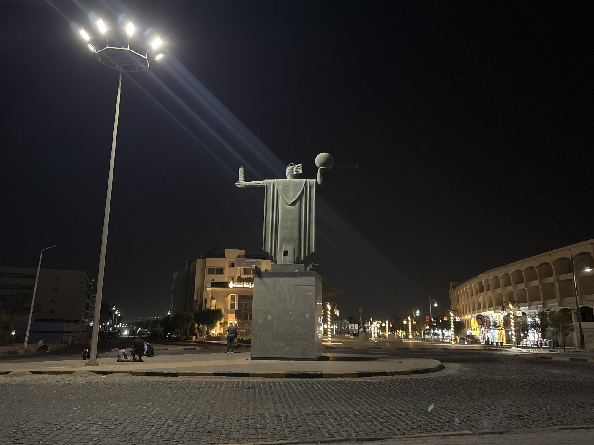 A nighttime scene shows a statue in the middle of a paved roundabout. The statue depicts a figure holding an object in each outstretched hand. A tall street light illuminates the statue and the surrounding area. In the background are buildings, including a large, arched structure, and several smaller lights along the street. People are present on the periphery, some standing near the statue and others working or sitting near the edge of the roundabout. The environment is dark with illuminated areas, creating a stark contrast.