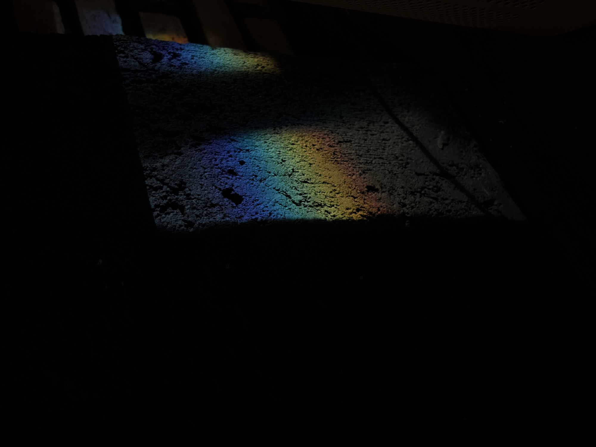 The image depicts a dark, textured wall in Nha Trang Ward, Vietnam, illuminated by a narrow band of refracted light that forms a rainbow spectrum. The light is cast from an unseen source, possibly a window or opening, and falls across the left side of the wall, creating a sharp diagonal contrast between shadow and color. The wall's surface is rough and uneven, with visible pores and irregularities. To the right of the wall, a darker area suggests the continuation of the environment, with faint horizontal lines hinting at a structural element or possibly another surface. The overall scene is minimalist, focusing on the interplay of light, shadow, and color on a distressed surface. There are no people or animals present.