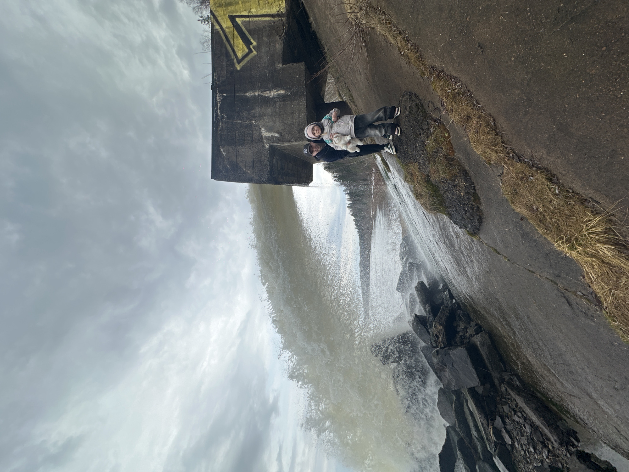 Two individuals, an adult and a child, are standing near a dam in Noviy, Russia. The child is holding a stuffed animal. Water is flowing over the dam, creating a waterfall effect. The sky is overcast. The area surrounding the dam appears to be concrete and stone.