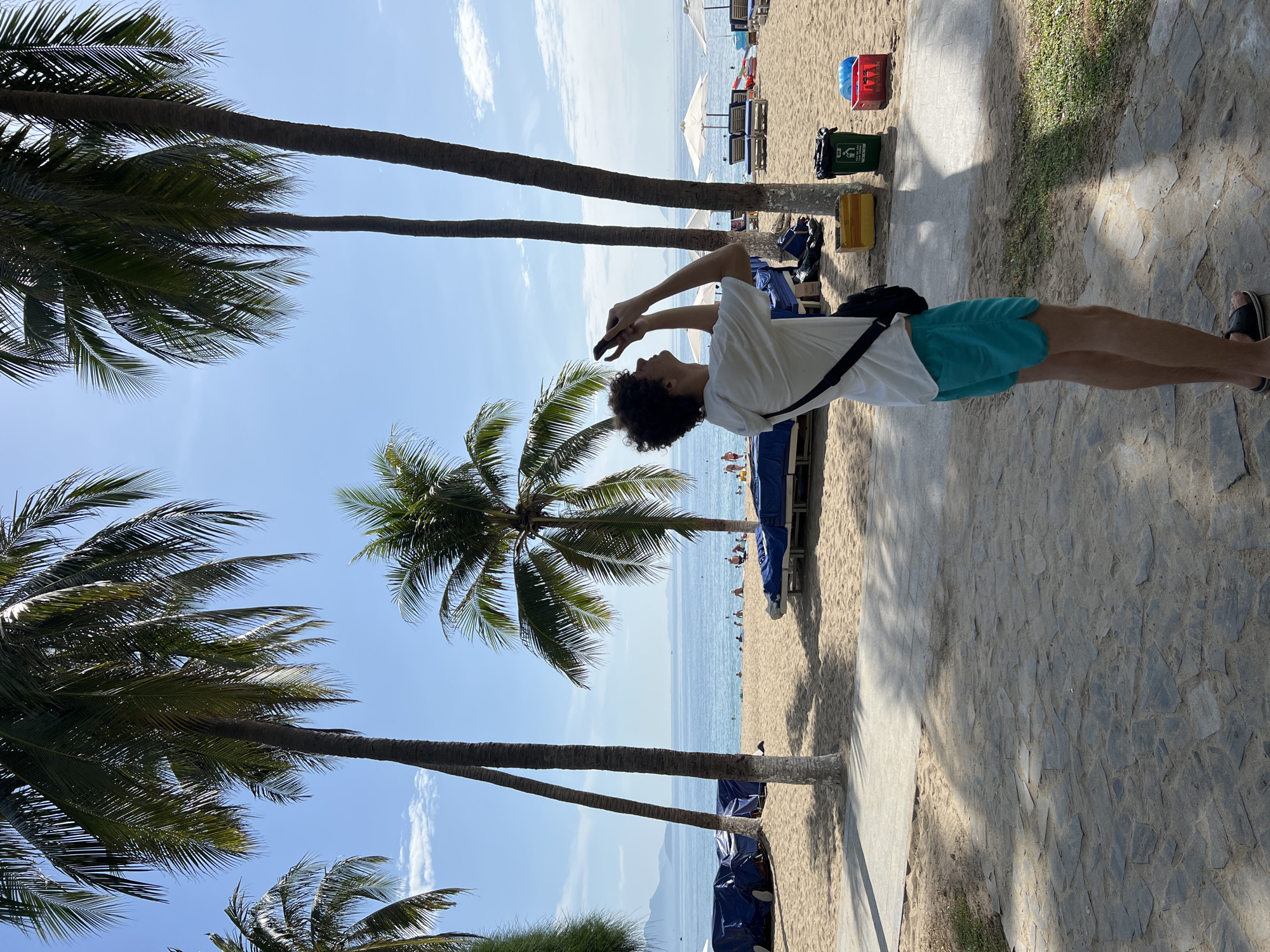 A young Caucasian male with curly brown hair, wearing a white short-sleeved hoodie over a t-shirt and turquoise swim trunks, stands on a stone pathway, looking up and holding a smartphone to take a photo. He has a black crossbody bag slung over his right shoulder. The background is a sandy beach in Nha Trang, Vietnam, with several palm trees casting shadows on the pathway. The ocean is visible beyond the beach, with a clear blue sky overhead. Several beach chairs with blue coverings, umbrellas, and some storage bins are present on the sand. There are other people visible in the distance on the beach and in the water.