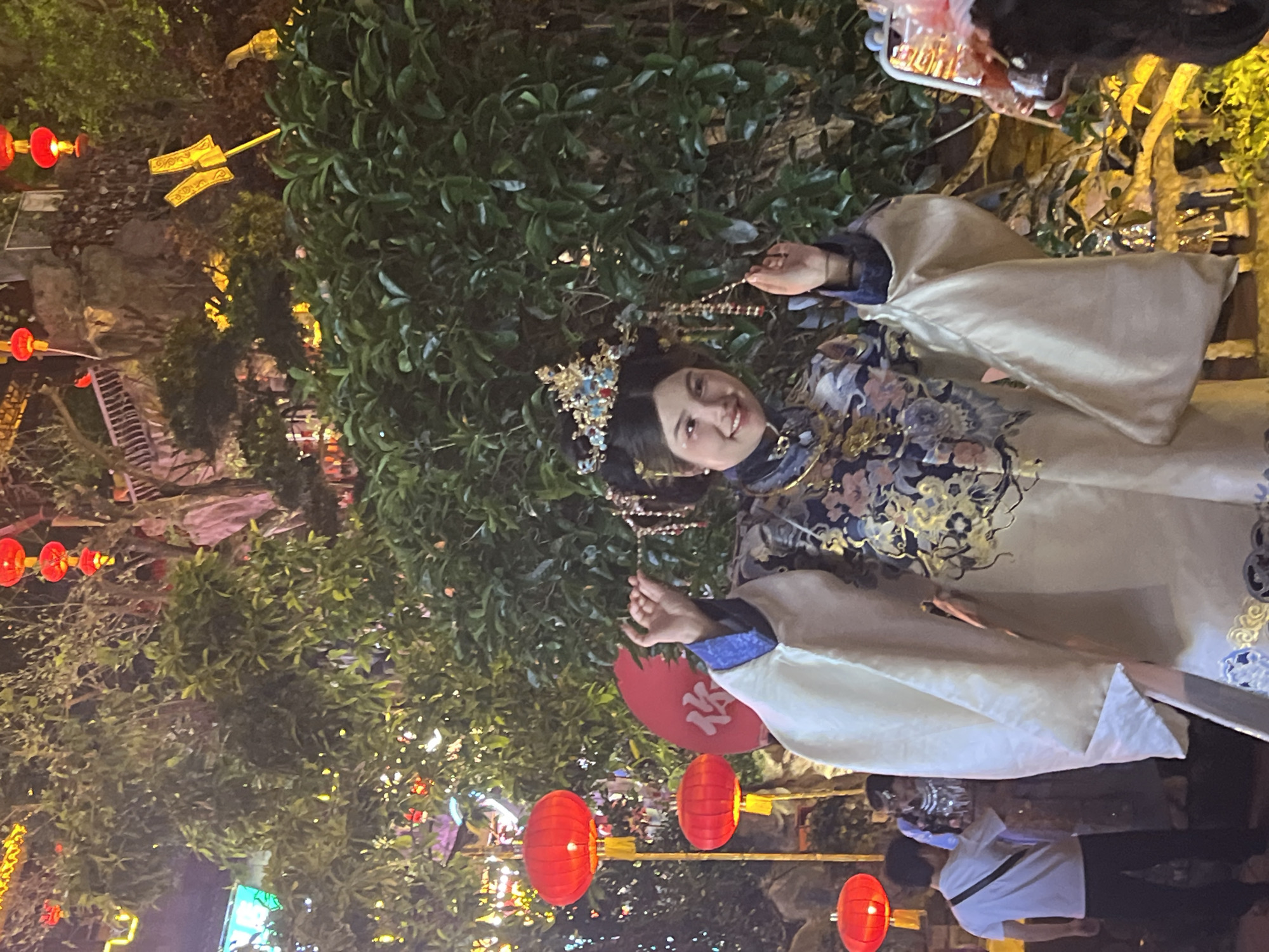 A woman in historical Chinese clothing smiles at the camera, adorned with an elaborate headpiece. She stands amidst a backdrop of greenery and traditional red lanterns. The location is the city of Yuzhong District, China. Other people and trees are also visible.