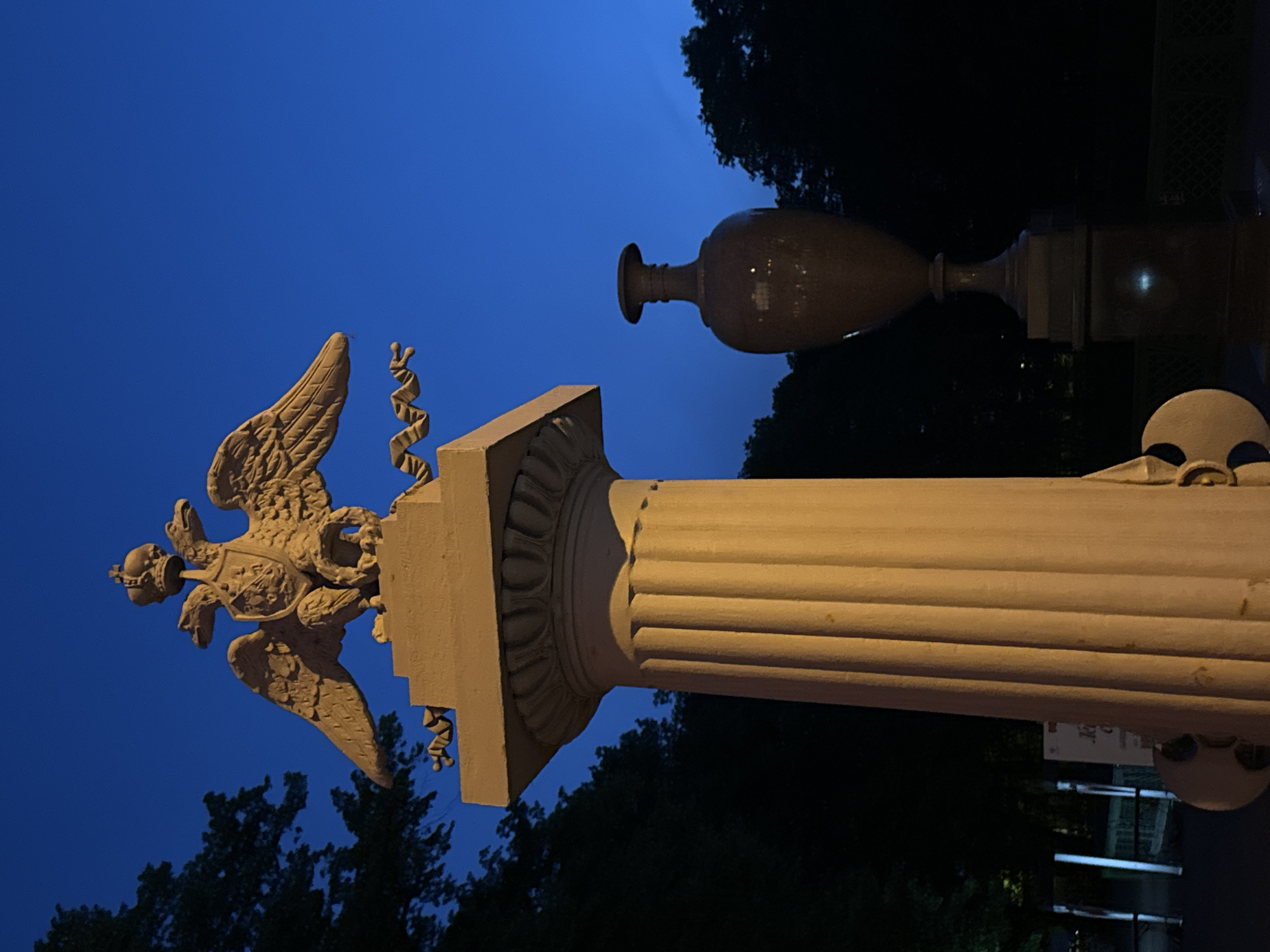 The image presents a detailed view of a stone monument within the Summer Garden in Saint Petersburg, Russia, observed during twilight or nighttime.
The primary subject is a cream-colored stone sculpture of the Russian Imperial double-headed eagle. The eagle is crowned, features two heads looking in opposite directions, and has a shield on its chest. It rests on a square plinth, which is supported by the decorative capital of a tall, fluted column of the same cream-colored stone. The column's shaft exhibits prominent vertical fluting, and its base is partially visible at the bottom of the frame. Ribbons or scrolls are depicted trailing from the eagle's feet.
To the right of the column, a darker, classical-style urn stands on a rectilinear pedestal. The urn has a distinct neck and a rounded body, with subtle reflections on its surface indicating a polished material or nearby light sources.
The background is dominated by a deep blue sky, characteristic of the blue hour. Dark, dense tree foliage is silhouetted against the sky, indicating a park or garden setting. In the lower left background, beyond the base of the column, indistinct luminous vertical elements and a faint signboard are visible, suggesting distant pathways or street furniture. No people are present in the scene.