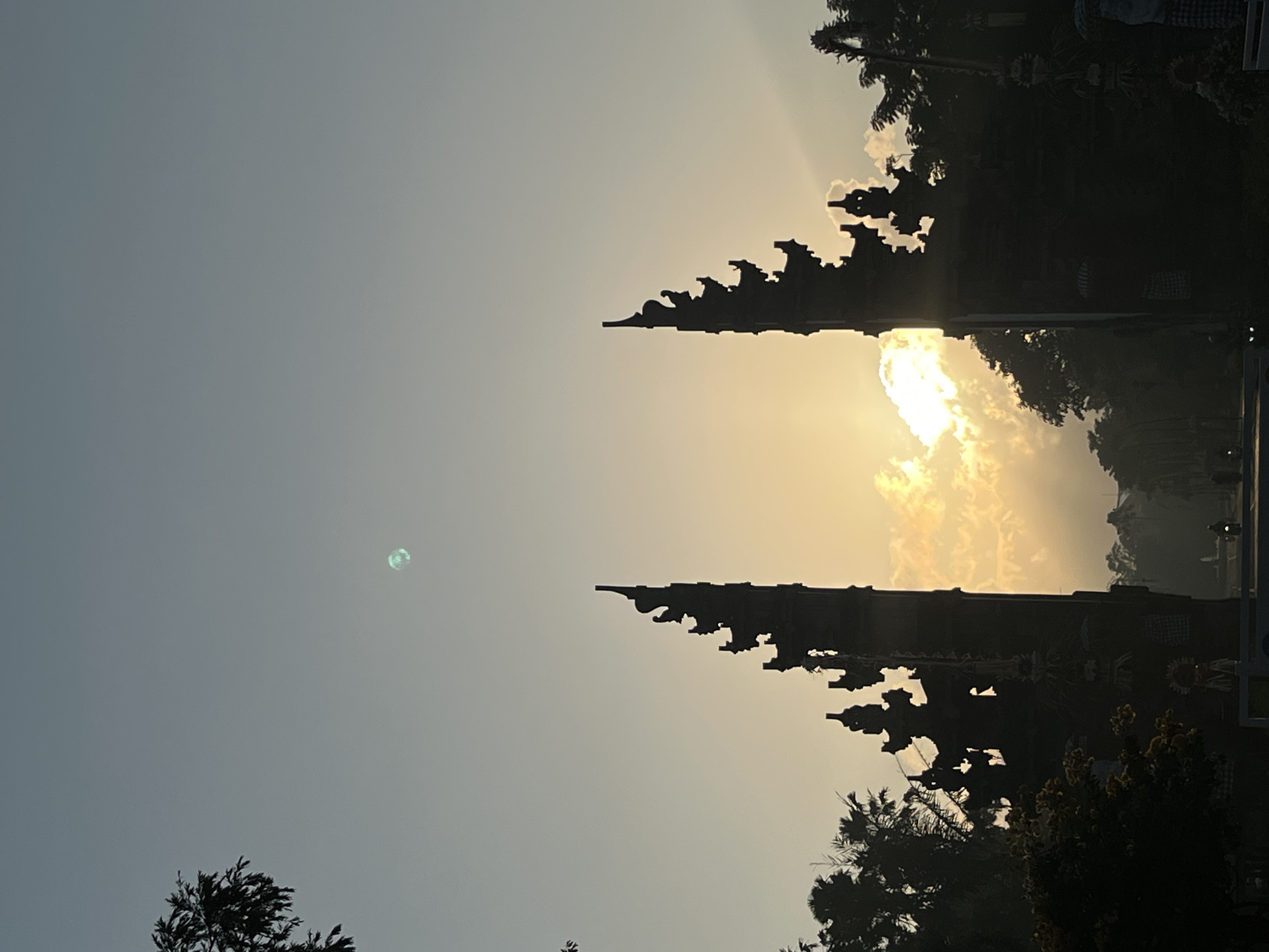 The image depicts a view through a traditional Balinese gate in Kuta Selatan, Indonesia, at sunrise. The gate structures, silhouetted against the bright sky, are intricately carved with tiered roofs and decorative elements. The sun is rising behind the gate, casting a warm glow through the clouds and illuminating the sky with hues of yellow and orange. Lush green trees and foliage frame the scene on either side of the gate, creating a tropical atmosphere. In the foreground, a street is visible with a single motorcycle rider in the distance, suggesting a peaceful morning in the area. The sky is a soft gradient of pale blue and yellow, with a faint, circular object that may be the moon visible in the upper portion of the frame.