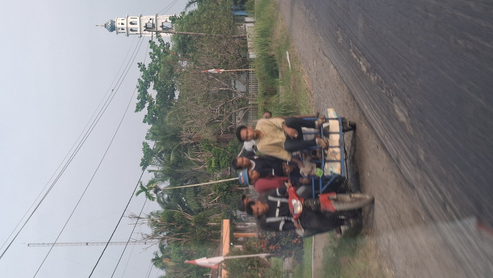 A group of six men are riding on a modified red motorcycle with a blue trailer attached. The motorcycle is being driven by a man in a black jacket with white stripes. Behind him, seated in the trailer, are five other men. One man in a light brown shirt is sitting in the front of the trailer, looking towards the camera. Another man wearing a red shirt and a blue hat is seated behind him. Two men in dark shirts are also in the trailer, and a younger man is seated in front of them. The motorcycle and trailer are on a dirt road next to an asphalt road. In the background, there is lush green vegetation, palm trees, a white fence, and a white tower with a blue dome, likely a mosque. A tall radio tower is visible in the distance to the left, and a flag is planted near a building on the left side of the image. The sky is overcast. This scene appears to be in Air Hitam, Indonesia.