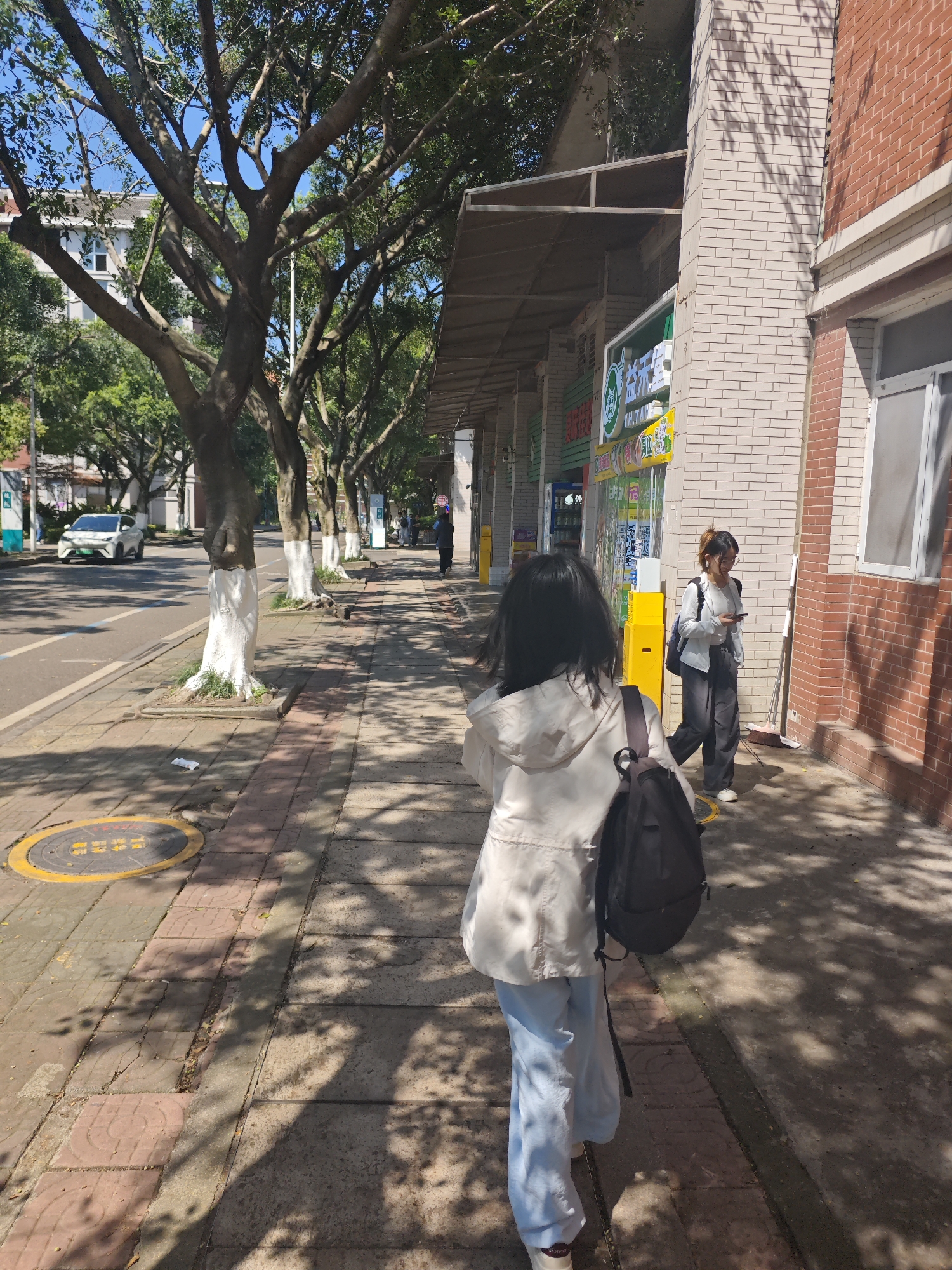 The image depicts a sunny street scene in Chongqing High-tech Zone, China. A young woman with a backpack, wearing a light-colored jacket and blue pants, is walking away from the camera on a sidewalk. Another young woman is walking in the opposite direction, looking at her phone. Trees line the street, their trunks painted white at the base. Buildings are adjacent to the sidewalk; a multi-story building is across the street. A white car is visible on the street. The sidewalk is paved with light-colored tiles. There are shops or businesses on the side of the sidewalk. A manhole cover is on the pavement near the street. Shadows from the trees dapple the sidewalk.