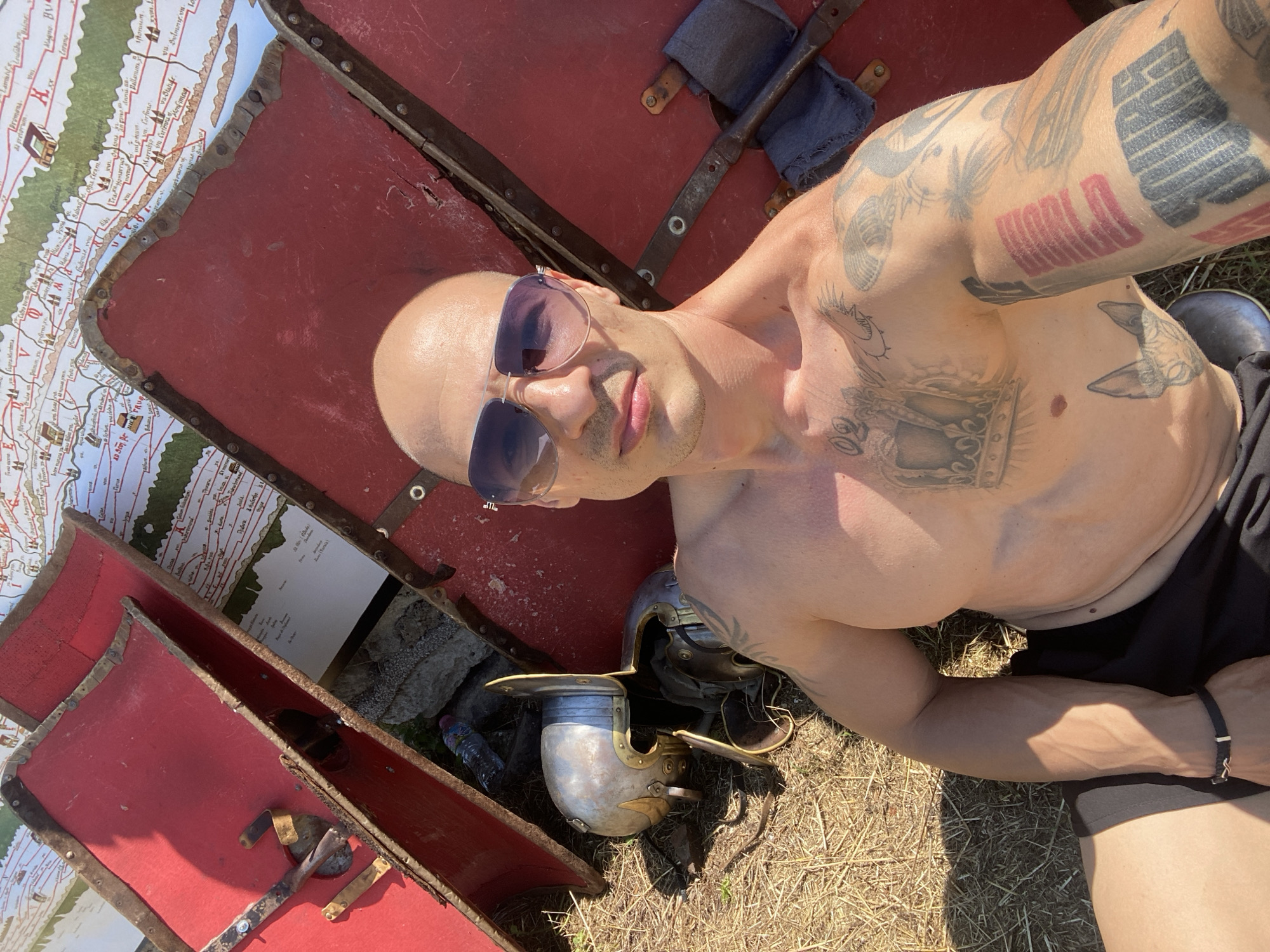 An outdoor selfie captures a bald male individual with a tanned complexion, wearing purple-tinted aviator sunglasses, black shorts, and a black and metallic bracelet on his left wrist. He is shirtless, prominently displaying a large crown tattoo with the number "02" on his left pectoral, a Sphynx cat head tattoo on his left abdomen, and various other tattoos on both arms, including abstract shapes and text. He is seated on dry, yellowed grass under bright sunlight, with distinct shadows visible.
To the subject's left, two Roman-style helmets, likely Galea, are resting on the ground alongside a partially visible clear plastic water bottle. Behind the individual and extending across the frame are multiple red, curved, rectangular Roman scutum shields, appearing worn with exposed wood and metal banding secured by rivets. A dark grey piece of fabric is draped over the handle of one shield on the right. In the background, behind the shields, a large, detailed map depicting geographical features and settlements, consistent with an ancient Roman style, is visible. The scene is situated in Gigen, Bulgaria, suggesting a historical reenactment or archaeological context. The individual is looking directly at the camera.