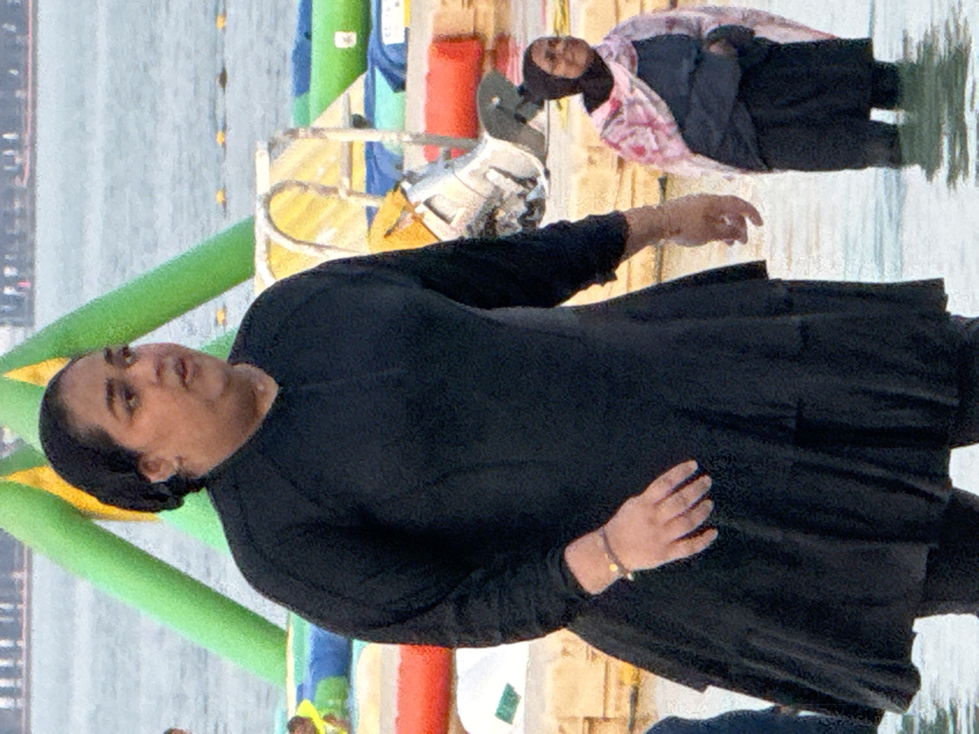 A medium shot captures two women wading in shallow, rippling water, with inflatable structures and a body of water in the background. The woman in the foreground, on the left, is facing right, wearing a black burkini that reaches her knees and has long sleeves. Her left arm is bent, and her hand, adorned with a bracelet, is resting on her hip. The woman on the right, in the background, is facing forward and slightly left, wearing a dark blue burkini and a pink and white patterned hijab. She is standing with her hands clasped in front of her. The water is a light gray, and the background features green and yellow inflatable structures, with a blurry, distant cityscape under a hazy sky. The lighting is natural, suggesting an outdoor, possibly daytime setting.