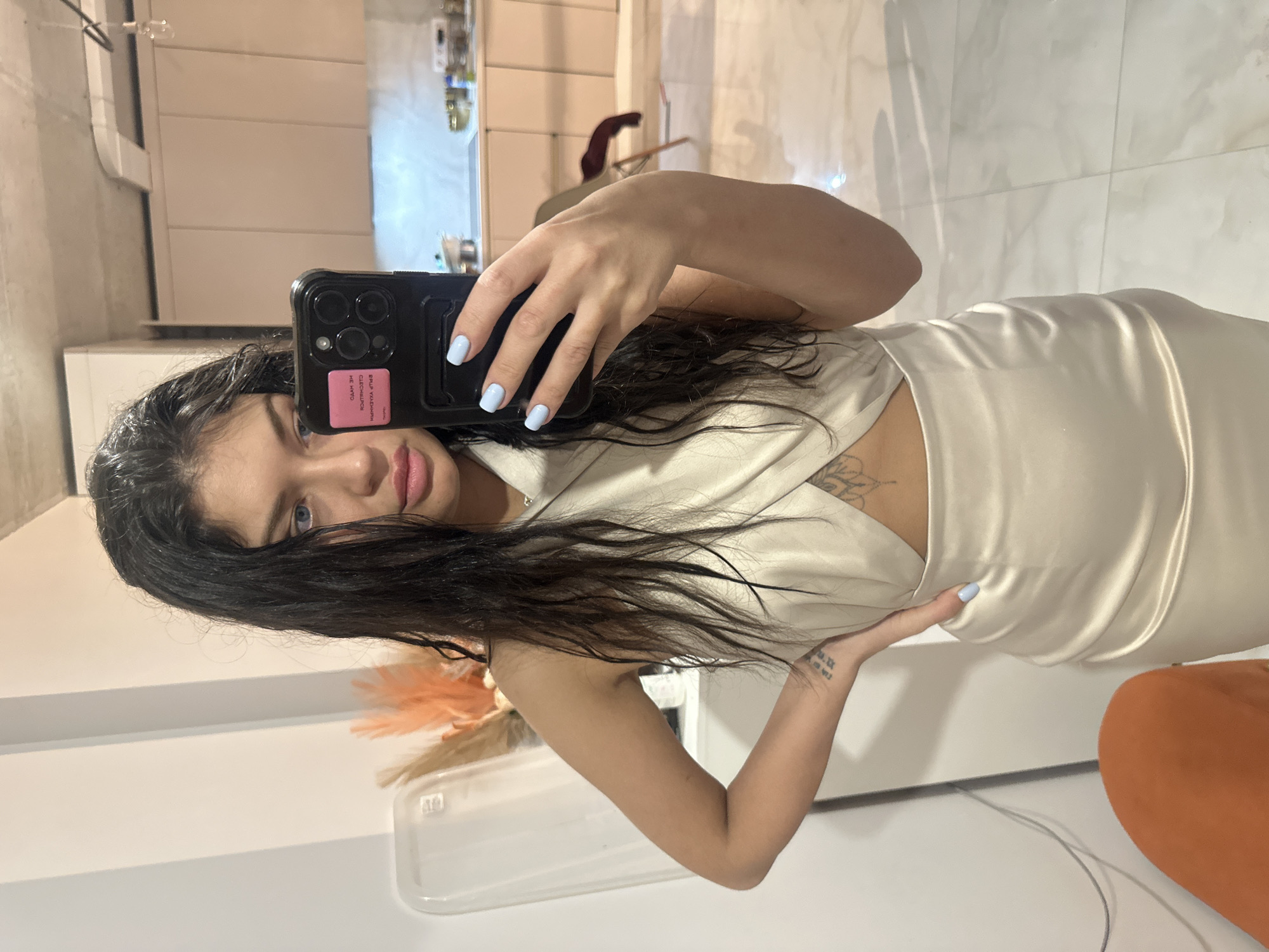 The image is a mirror selfie of a young woman, rotated 90 degrees counter-clockwise, taken indoors in what appears to be a modern living space in Khimki, Russia. The woman has long, dark, wavy hair and light blue eyes, looking directly at the camera with a neutral expression. She wears a light-colored, possibly off-white or cream, satin-like halter neck dress with a midriff cut-out.
She holds a dark-colored smartphone, likely an iPhone, in her right hand to capture the reflection. Her fingernails are painted light blue. A pink rectangular sticker on the phone case displays partially visible Cyrillic text, which seems to include "ОЛЕНО" and "КИМЧУК".
Tattoos are visible on her body: a leafy or floral design on her upper left chest/shoulder area, and Roman numerals "VI.IX. MM.XXI" (representing June 9, 2021) on her left forearm.
The reflected background shows modern interior elements, including white tiled walls or flooring, an exposed ventilation duct or pipe, and a dark chair with a reddish cushion. An orange decorative element, possibly dried pampas grass, and a clear plastic container sit on a surface. A large orange, soft object, likely a cushion or furniture piece, is also visible in the lower right corner. The lighting suggests an indoor setting with artificial illumination.