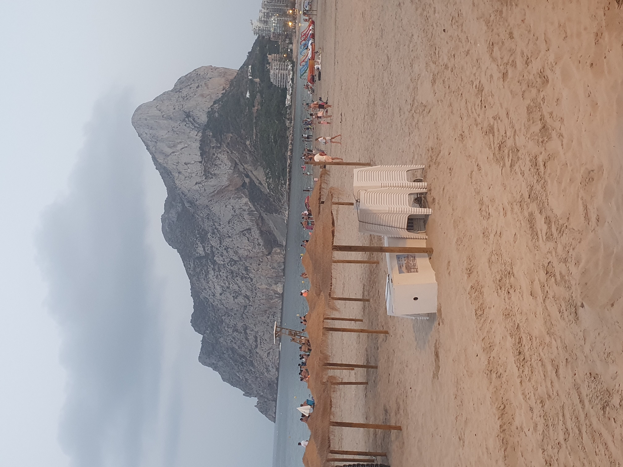 The image shows a beach in Calp, Spain, with the iconic Penyal d'Ifac rock formation in the background. The sky is overcast, and the lighting suggests it is late afternoon or early evening. Several straw umbrellas are set up on the sand, and stacks of white plastic chairs are visible in the foreground. A white cooler or service cart is also present. The sea is calm, and there are numerous people on the beach, some swimming and others relaxing. In the distance, there are buildings lining the coast, suggesting an urban area. There are also inflatable water toys on the beach near the water.