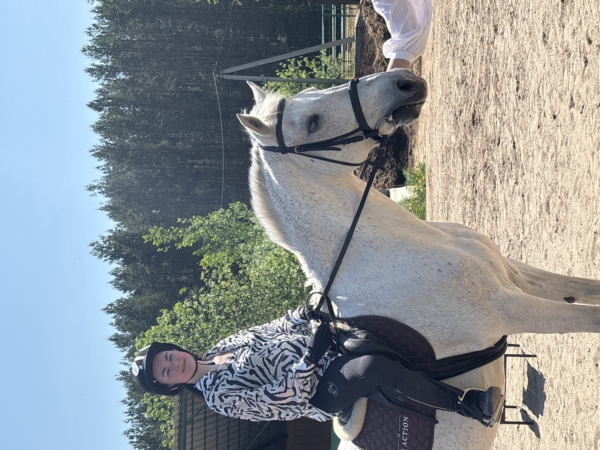 A light-skinned person with dark hair is seated on a white horse in an outdoor setting. The person wears a black riding helmet, a zebra-patterned shirt, black riding pants, and black riding boots. They are holding the reins. The horse is equipped with a black bridle, saddle, and girth. The background includes trees, a blue sky, and sandy ground. Part of a building is visible. There is an individual visible behind and to the right of the horse, wearing white. The location is likely a riding area, though a city or precise location in Русский Вожой, Russia, cannot be confirmed based on the image.