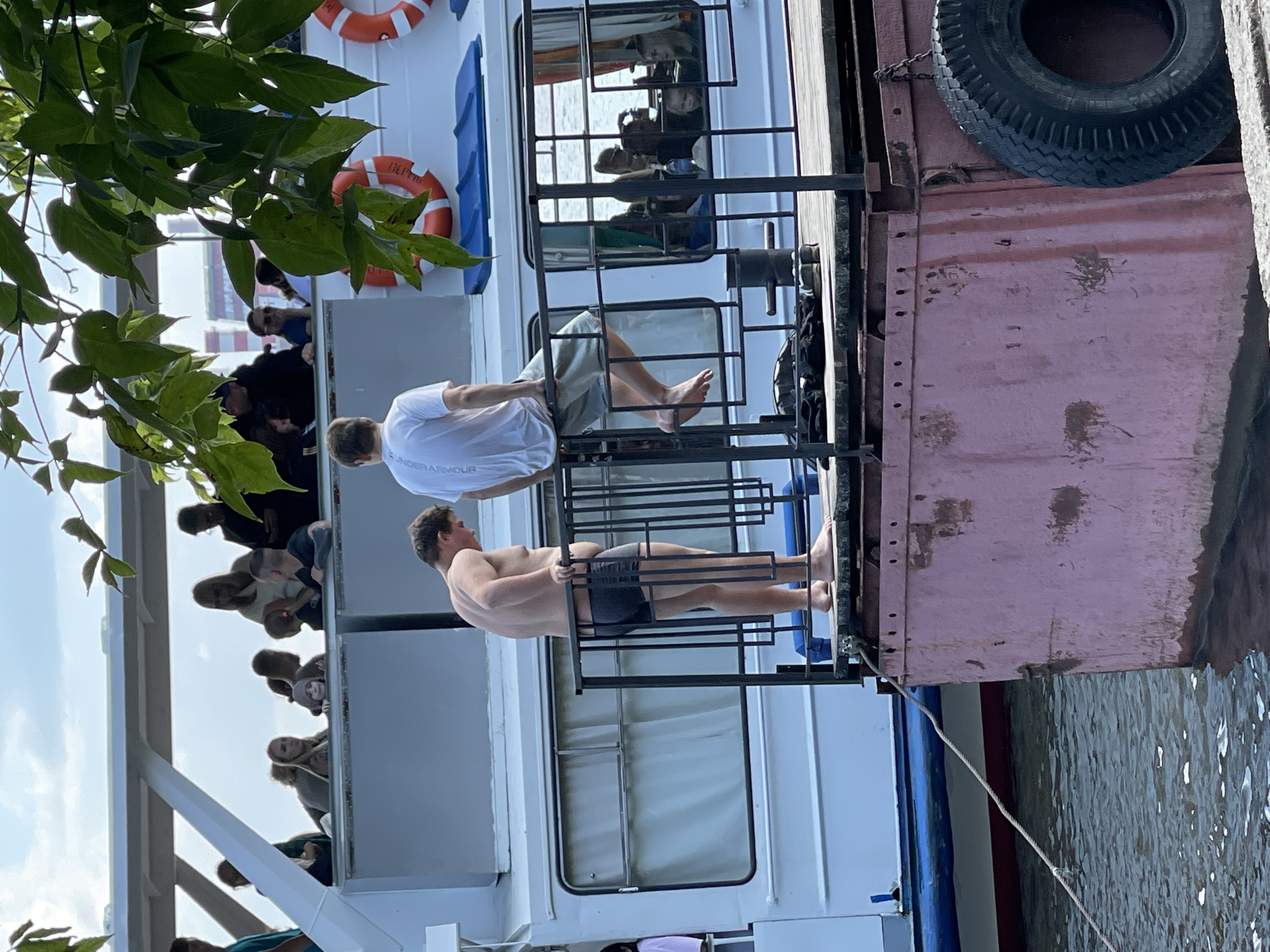 The image shows a close-up view of a section of a boat or floating structure, with two young men standing on a wooden deck. The man on the left is shirtless and wearing dark swim trunks, facing away from the camera and looking down at the water. He has a medium build. The man on the right is wearing a white t-shirt and light-colored shorts, sitting on a railing with his legs dangling over the side. He is also facing away from the camera. Behind them, people can be seen on the upper deck of a larger vessel, some looking out over the railing. The water is visible in the foreground, and a portion of a pink-colored structure with a large tire attached to its side is in the lower right of the frame. The scene appears to be on a warm, overcast day, with some green foliage in the upper right corner. The context suggests this is a public area in Izhevsk, Russia, likely a waterfront or river excursion.