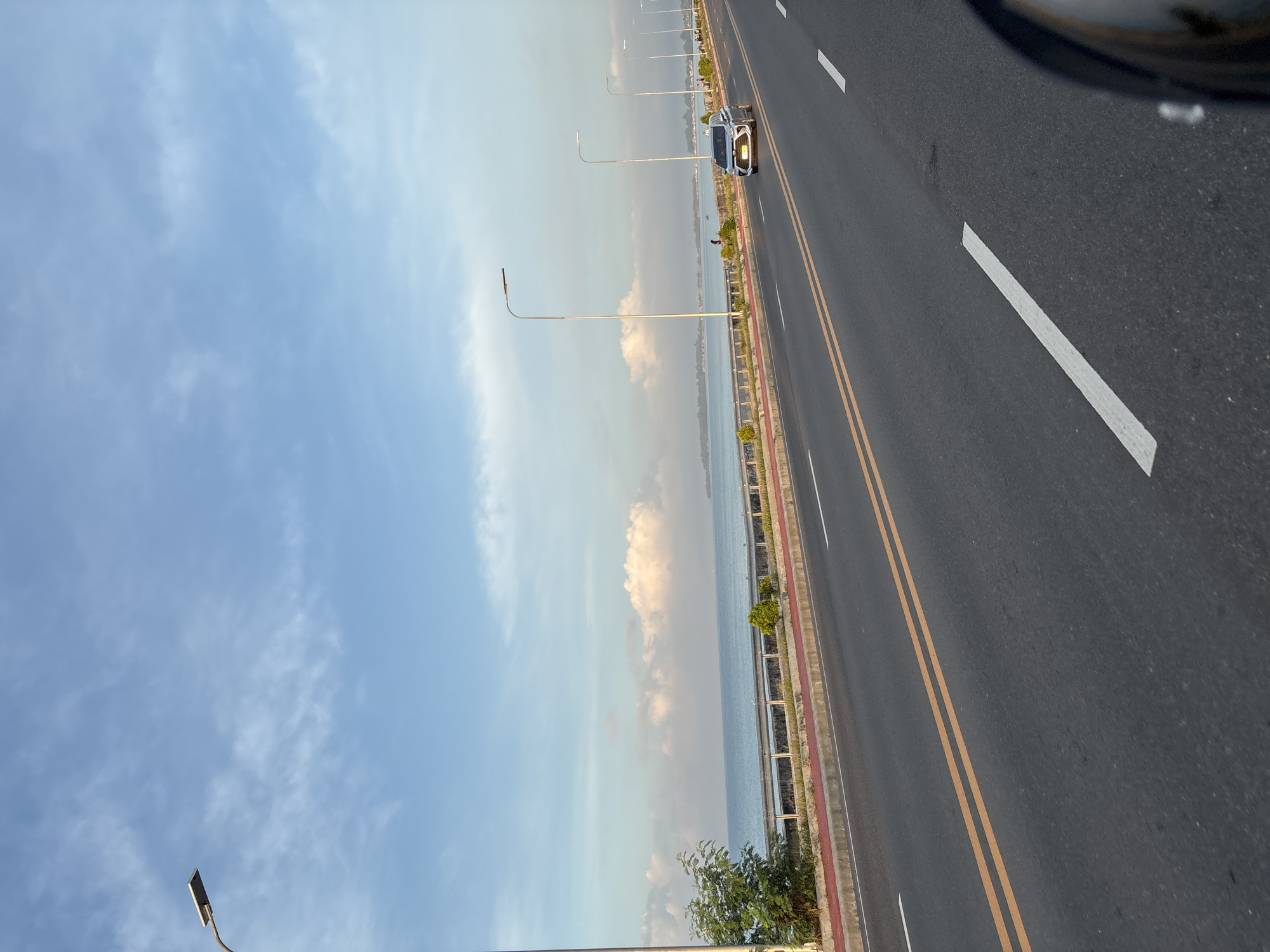 The image depicts a coastal road in Davao City, Philippines, likely along the Davao Gulf. The scene is captured from a moving vehicle, suggesting a journey along the highway. The road itself is paved with asphalt and marked with double yellow lines and white lane dividers. A silver SUV is traveling in the right lane, while other vehicles are not clearly visible in the distance.
To the left of the road, a sidewalk with a red border is present, lined with sparse vegetation and trees. Beyond the sidewalk is a metal guardrail, separating the road from the sea. The Davao Gulf stretches out, calm and blue, with a hazy horizon where land is faintly visible. Scattered white clouds dot the clear blue sky. Several modern streetlights, a characteristic of well-developed roads, are positioned along the roadside, indicating the presence of infrastructure. The overall atmosphere is peaceful, with the gentle sea and sky creating a serene backdrop for the urban commute.