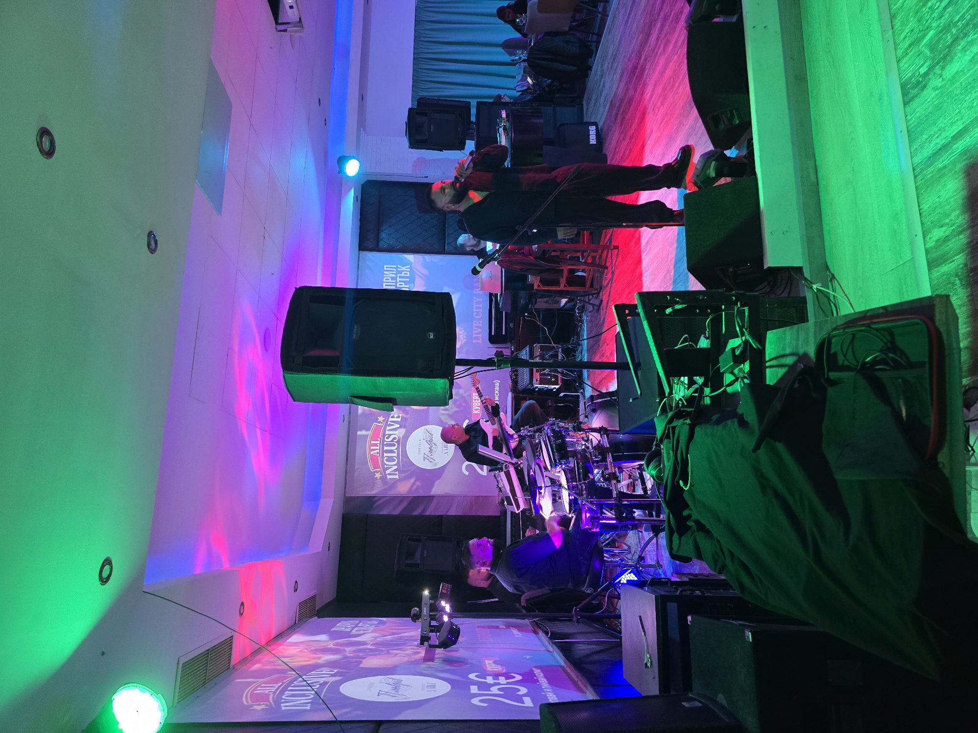 The image depicts a live musical performance at a venue with a stage, lighting, and projection screens. There are musicians on stage playing instruments, including a singer, guitarist, keyboard player, and drummer. The stage is illuminated with vibrant green, blue, and pink lighting, and the walls are also bathed in colored lights. Projection screens display the text "ALL INCLUSIVE" and "LIVE CITY RAN" along with other promotional material. Speakers and other sound equipment are visible around the stage. The OCR indicates the location of the performance as "LIVE CLUB" in "Пловдив" (Plovdiv), a city in Bulgaria. The overall setting suggests a lively event with a focus on music and entertainment.