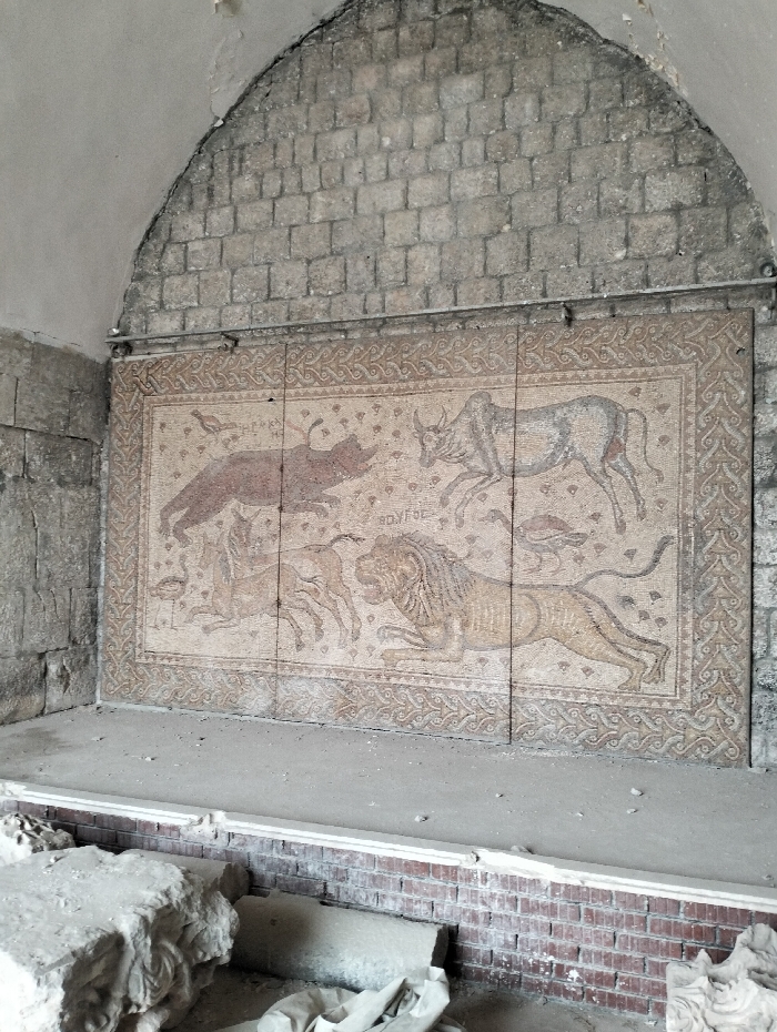 A mosaic featuring animal figures adorns a wall constructed of pale bricks. A boar, bull, and lion, along with other fauna, are depicted in shades of brown, grey, and beige. The mosaic has a decorative border and appears to be composed of smaller tiles. The wall is recessed under an arched, brick ceiling. Below the mosaic is a low brick wall with scattered rubble.