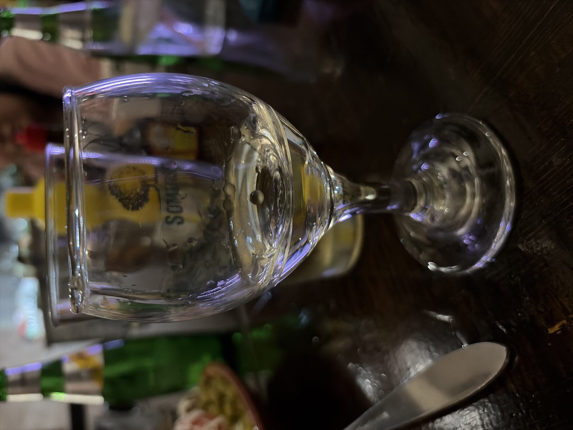 A clear glass wine glass with condensation sits on a dark, possibly wooden table. Visible through the glass is a yellow object with the partial word "SOMOS" written on it, possibly a bottle label. To the right of the glass is a silver knife, partially in focus. Other blurred objects, including green bottles and what appears to be food on a plate, are present in the background. The image suggests an indoor setting, possibly a restaurant or bar. There's no indication this image was taken in Galabovo, Bulgaria.