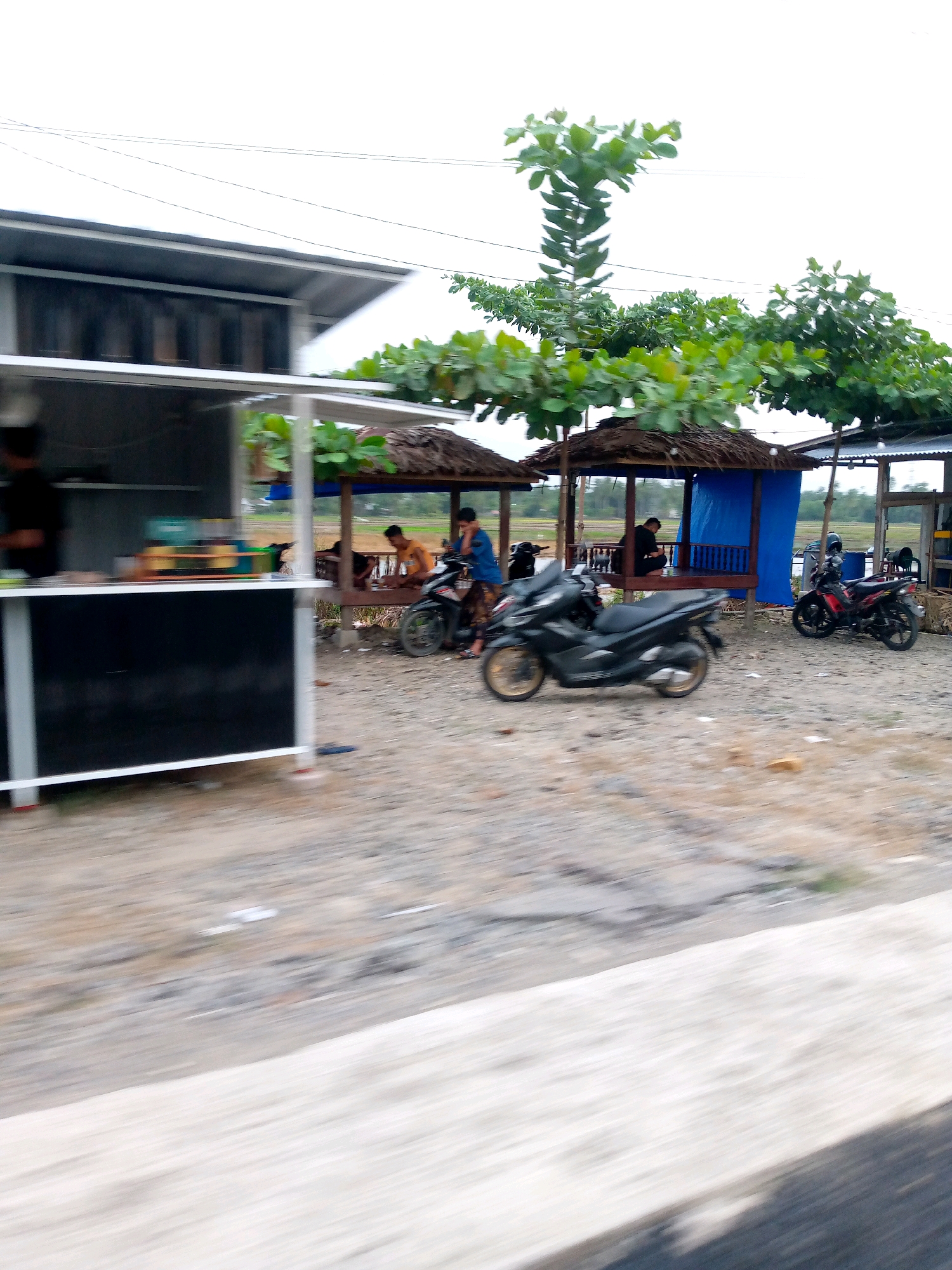 The image depicts a roadside scene in Keudeteupinpunti, Indonesia. In the foreground, a blurred motorcycle is prominently displayed. To the left, a white food stall with a black counter is visible. Beyond the stall, a thatched-roof gazebo with blue tarp walls provides shade. Two men are seated inside the gazebo, and another man is standing. Two motorcycles are parked near the gazebo: a black scooter-like motorcycle to the left and a red and black motorcycle to the right. The ground is covered with gravel and sand. In the background, trees with lush green foliage are present, and a power line stretches across the sky. The overall environment suggests a rural or semi-urban setting with small businesses catering to travelers.