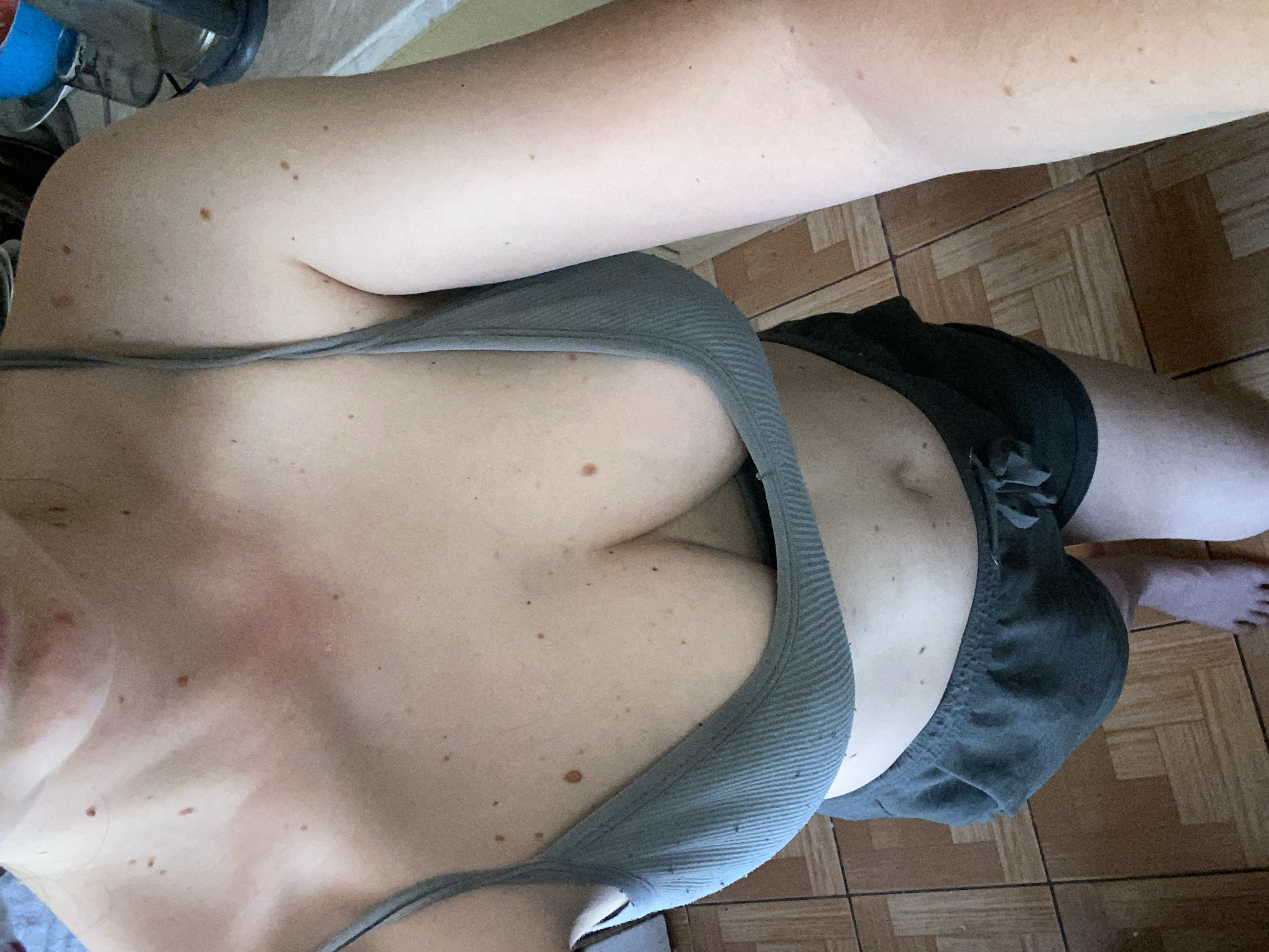 A person is shown from the chest down, wearing a gray bralette and dark gray shorts. They are standing on a floor with brown and tan square tiles. The person's skin is fair and has several moles visible on their chest, arms, and stomach. Their feet are bare. In the background, a portion of a countertop and some kitchen appliances are visible. The scene appears to be indoors.