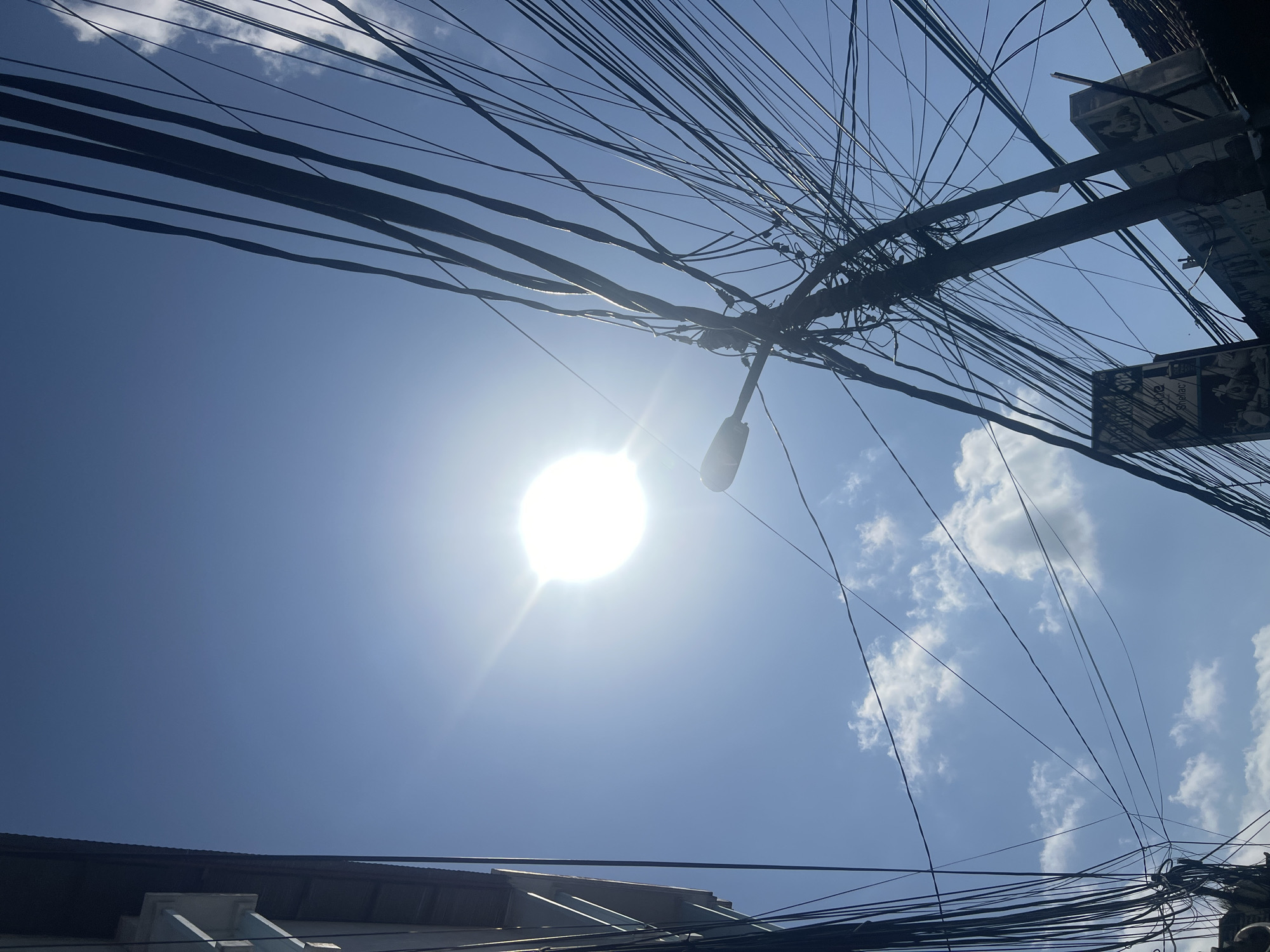 The image depicts a low-angle view looking up at a bright, hazy sun in a clear blue sky with scattered white clouds. A dense network of tangled black electrical wires dominates the right side of the frame, emanating from a utility pole that supports a streetlamp. On the left, the dark facade of a building is visible, with some architectural details suggesting a rooftop overhang. The ground level of the scene is hinted at by the presence of a sign attached to the utility pole, featuring text and images, partially obscured by the wires. The overall impression is that of a busy urban environment, likely a street in Kuta, Indonesia, characterized by overhead utility infrastructure.