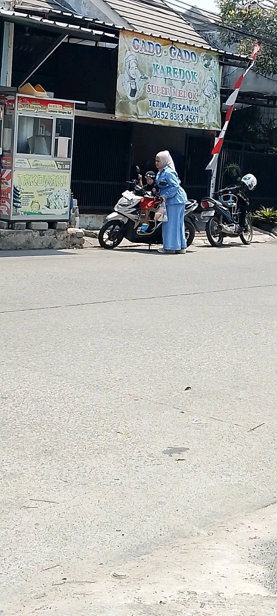 In Satriamekar, Indonesia, on a sunny day, a woman in a light blue hijab and matching outfit stands next to a white scooter parked on the side of a paved road. A young child, wearing a black headband and red shorts, is seated on the scooter. The woman is holding a white bag and appears to be interacting with the child. Behind them, a banner hangs from a building, displaying "CADO-CADO KAREDUK SUPER MEDOK TERIMA PESANAN 0852 8383 4567" along with an illustration of a woman in a hijab. To the left of the banner is a food cart with the text "TAKONIAKI" and pricing information. A second, darker colored scooter is parked further down the road to the right. The environment suggests a commercial area with buildings and a street.