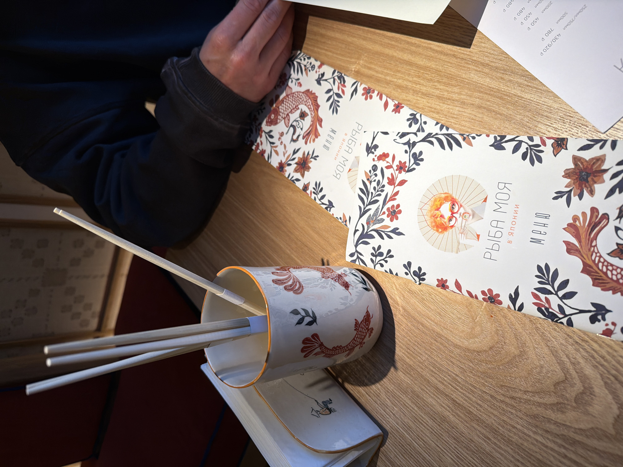 Here's a matter-of-fact description of the image:
A photograph taken at eye level depicts a table setting, likely in a restaurant. A person with a dark, cuffed jacket is visible on the left side, with their hand partially in frame as if reading a menu. Two menus with a decorative design featuring flowers and koi fish are visible, with the text "РЫБА МОЯ в Японии меню", indicating "My Fish in Japan menu" in Russian. A cup decorated with koi fish contains several chopsticks. The cup rests on a white dish or small book, adding to the table setting. The table is a light wood color, likely a laminate or veneer. An additional menu with prices is visible in the background. This scene is consistent with a Japanese restaurant, possibly located in Moscow, Russia, based on the Russian text on the menus.