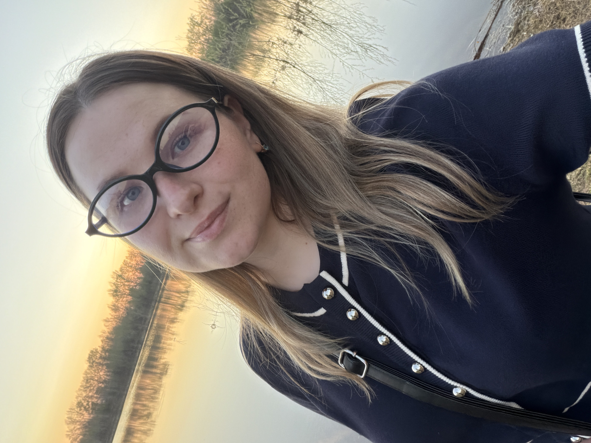 A woman with blonde hair and glasses stands near a body of water, possibly a lake or reservoir. She is wearing a dark blue cardigan with white trim and silver buttons, and a black crossbody bag. The background shows trees and a landscape illuminated by a warm, golden light, suggesting it is either sunrise or sunset. The location is Chaikovsky, Russia.