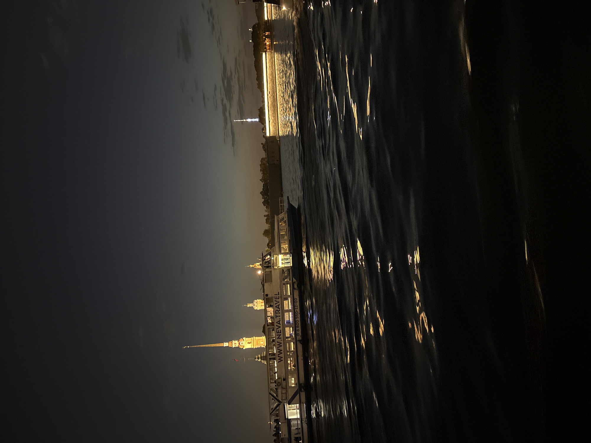 The image shows a nighttime view of Saint Petersburg, Russia, from a boat on the Neva River. Illuminated historical buildings and structures line the riverbank, including the Peter and Paul Fortress with its distinctive spire. A passenger boat with lit windows and the website "www.neva.travel" is visible, reflecting light onto the water's surface. The water's surface is rippled, creating distorted reflections of the city lights. A bridge is visible in the background. The sky is dark, indicating either early evening or late night.