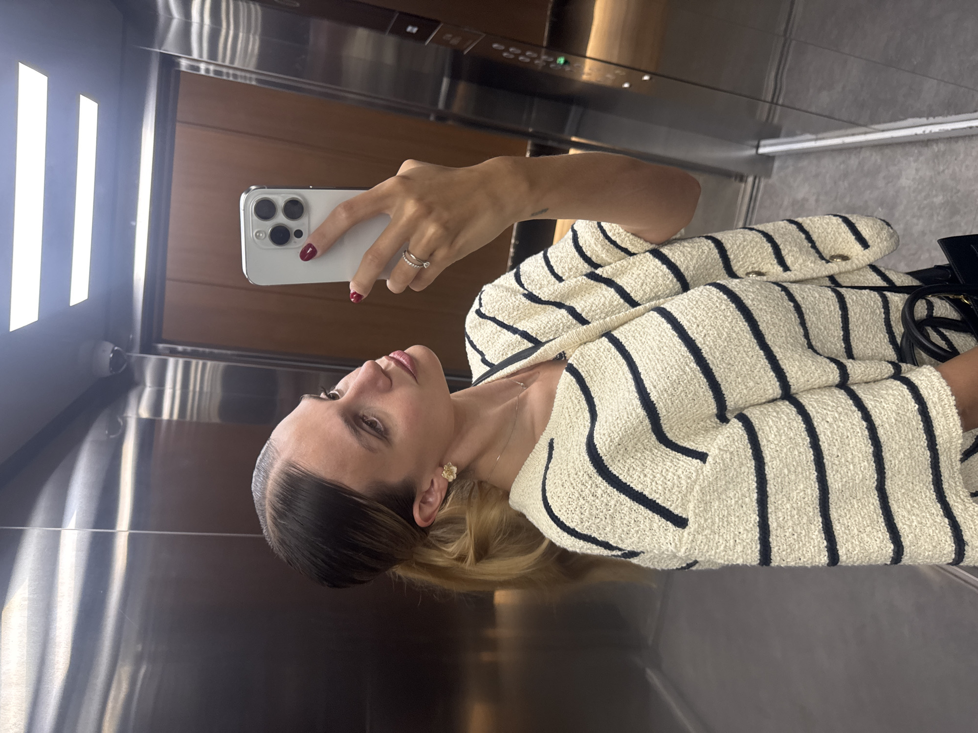 A young woman with blonde hair pulled back in a ponytail and wearing a cream-colored sweater with navy stripes takes a selfie in an elevator. She is wearing a delicate necklace and flower-shaped earrings, and has red nail polish. A ring with a small diamond is visible on her left hand, which is holding a smartphone. The elevator has stainless steel walls and wooden doors. The lighting appears to be overhead fluorescent lights.